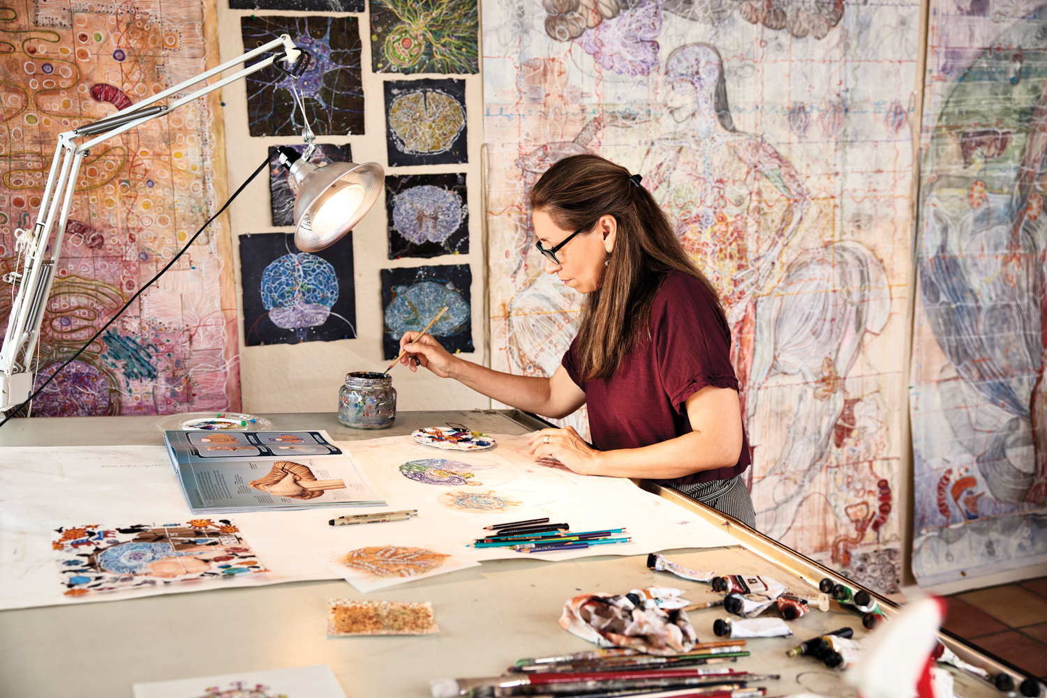 woman painting at a table in a studio surrounded by artwork