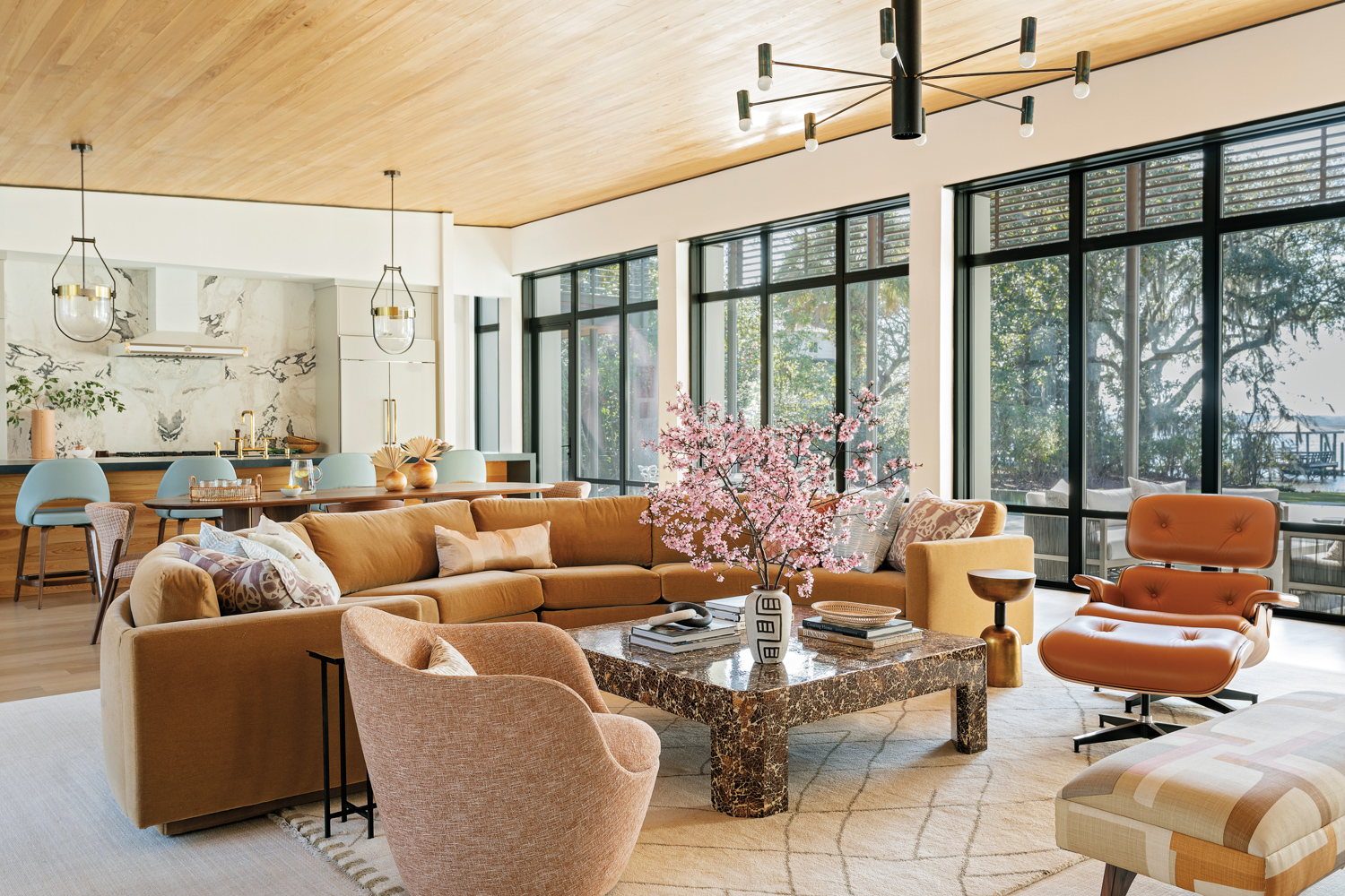 An open-format living room with warm-toned upholstery, a patterned rug, modern chandelier and vintage coffee table