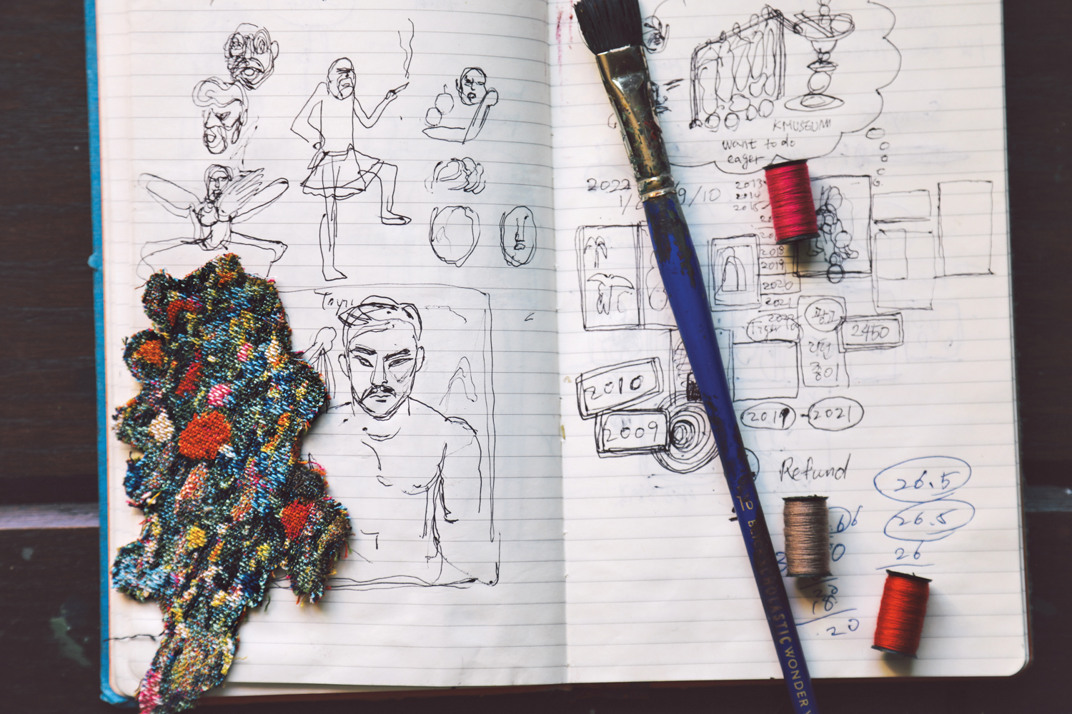 Pencil sketches in a notebook, with a patch of embroidery, a paintbrush and thread spools on the pages.