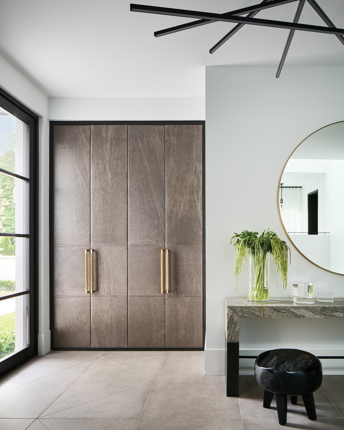 entry with leather-upholstered closet doors, marble console, black stool and circular mirror