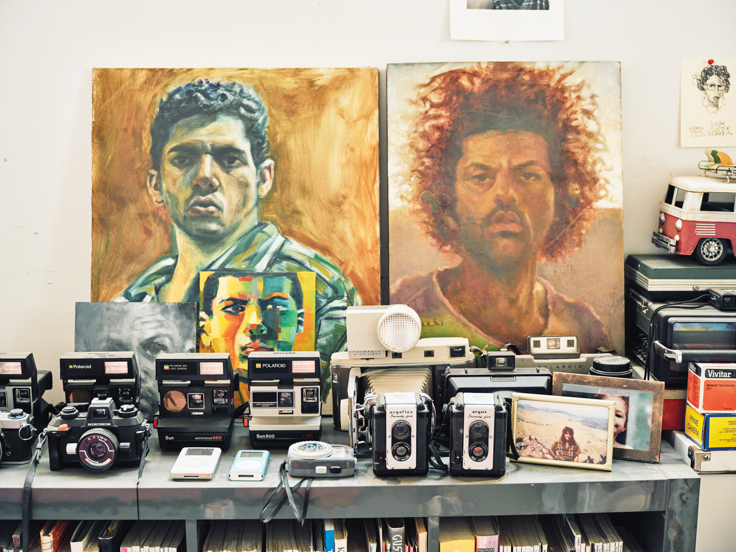 Two painted portraits by Michael Gadlin over a collection of vintage cameras.