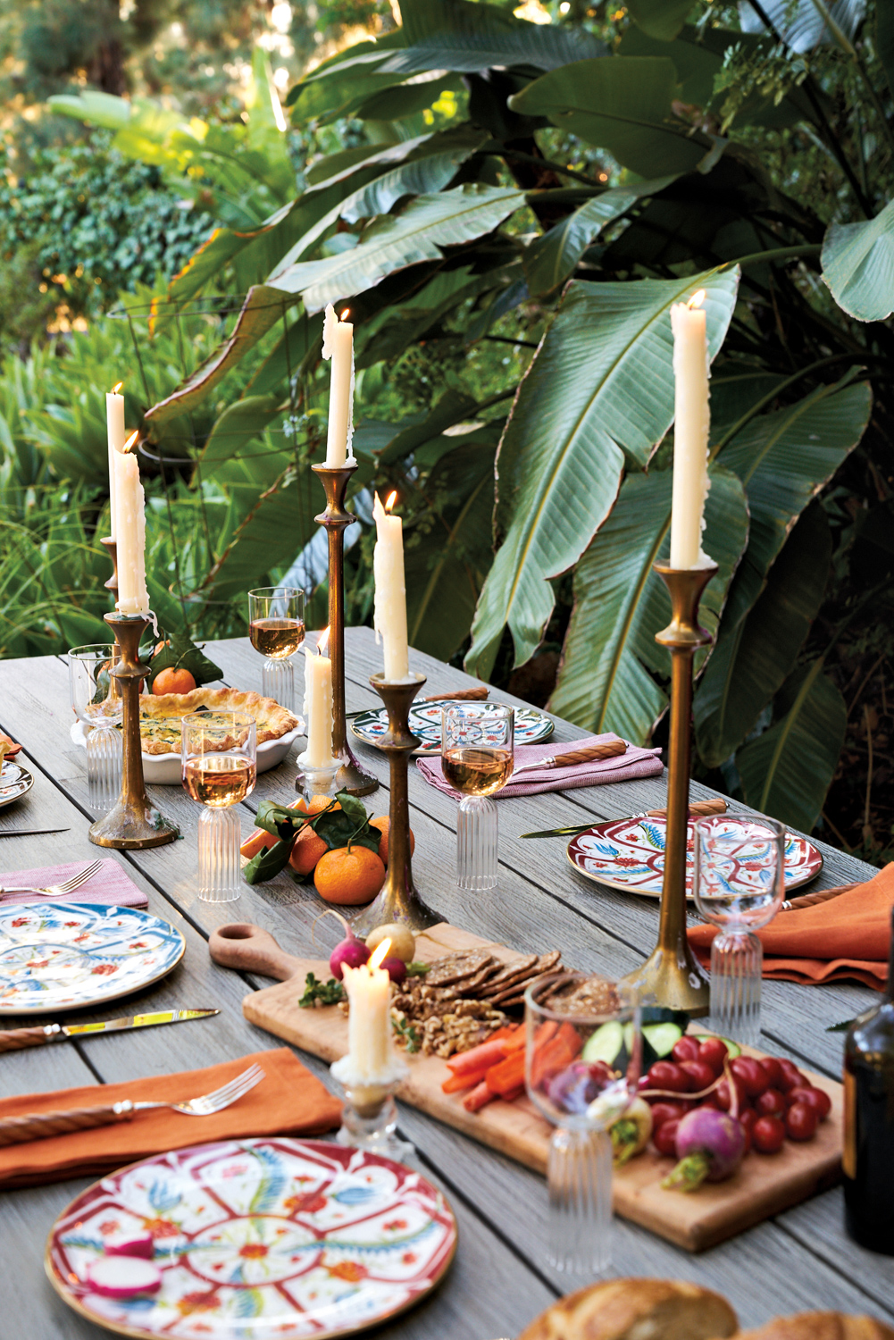 Outdoor table setting by Collecto with mismatched-patterned plates, cheese board and white taper candles in tall holders 