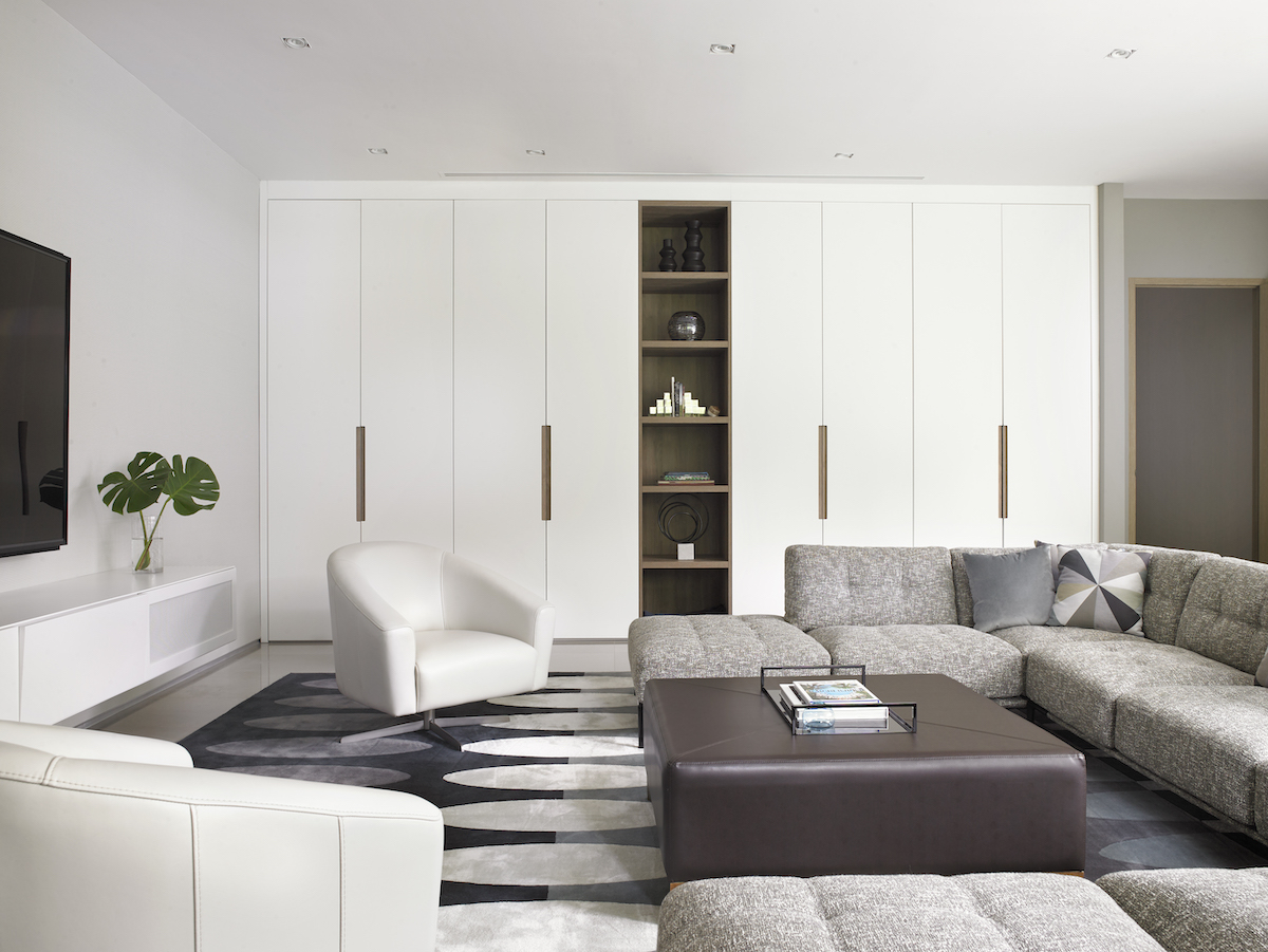 White wall of cabinets, geometric area rug, brown ottoman, grey sectional, white side chairs.