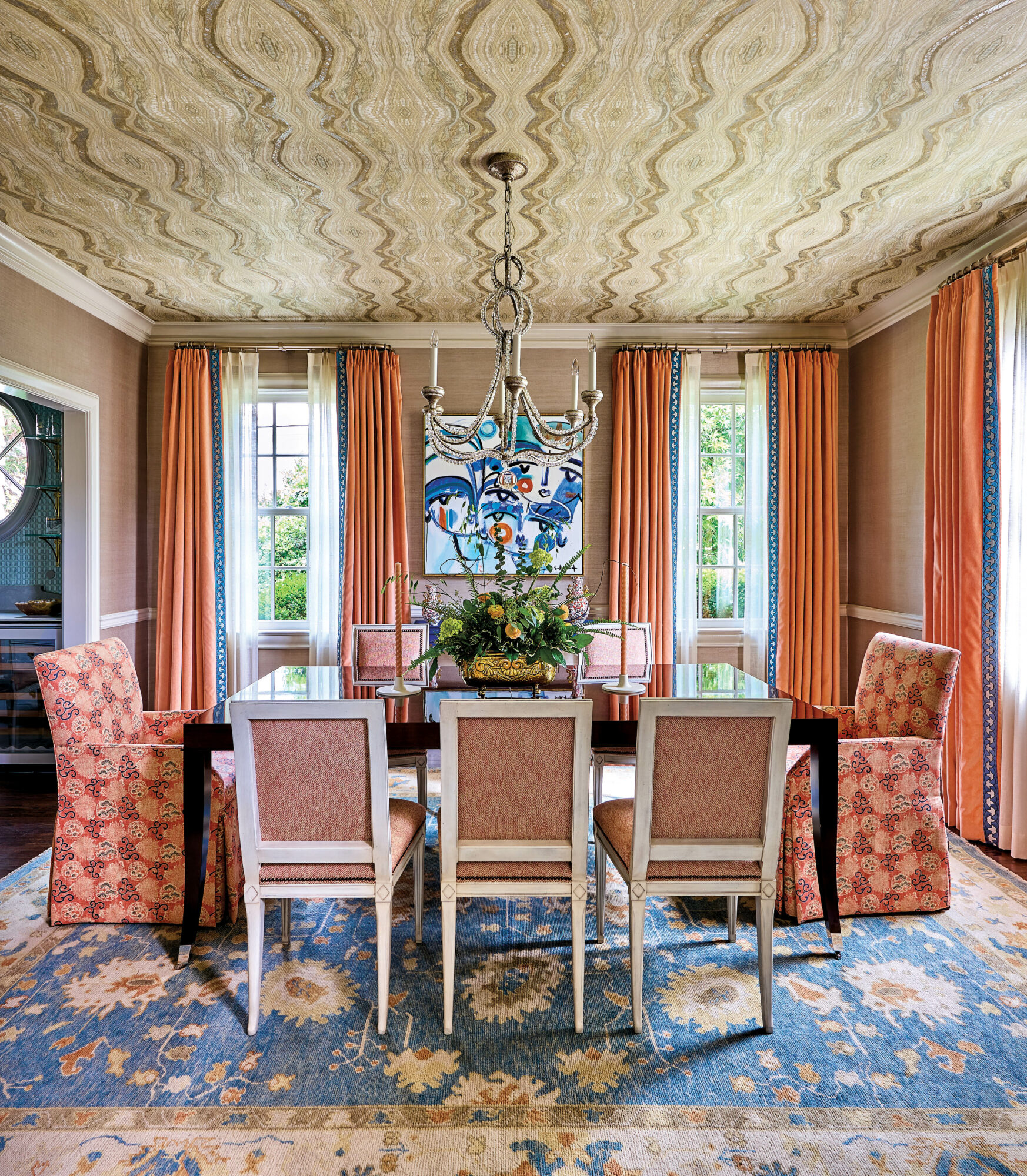 Dining room with coral fabrics, blue floral rug, abstract artwork and wallpapered ceiling