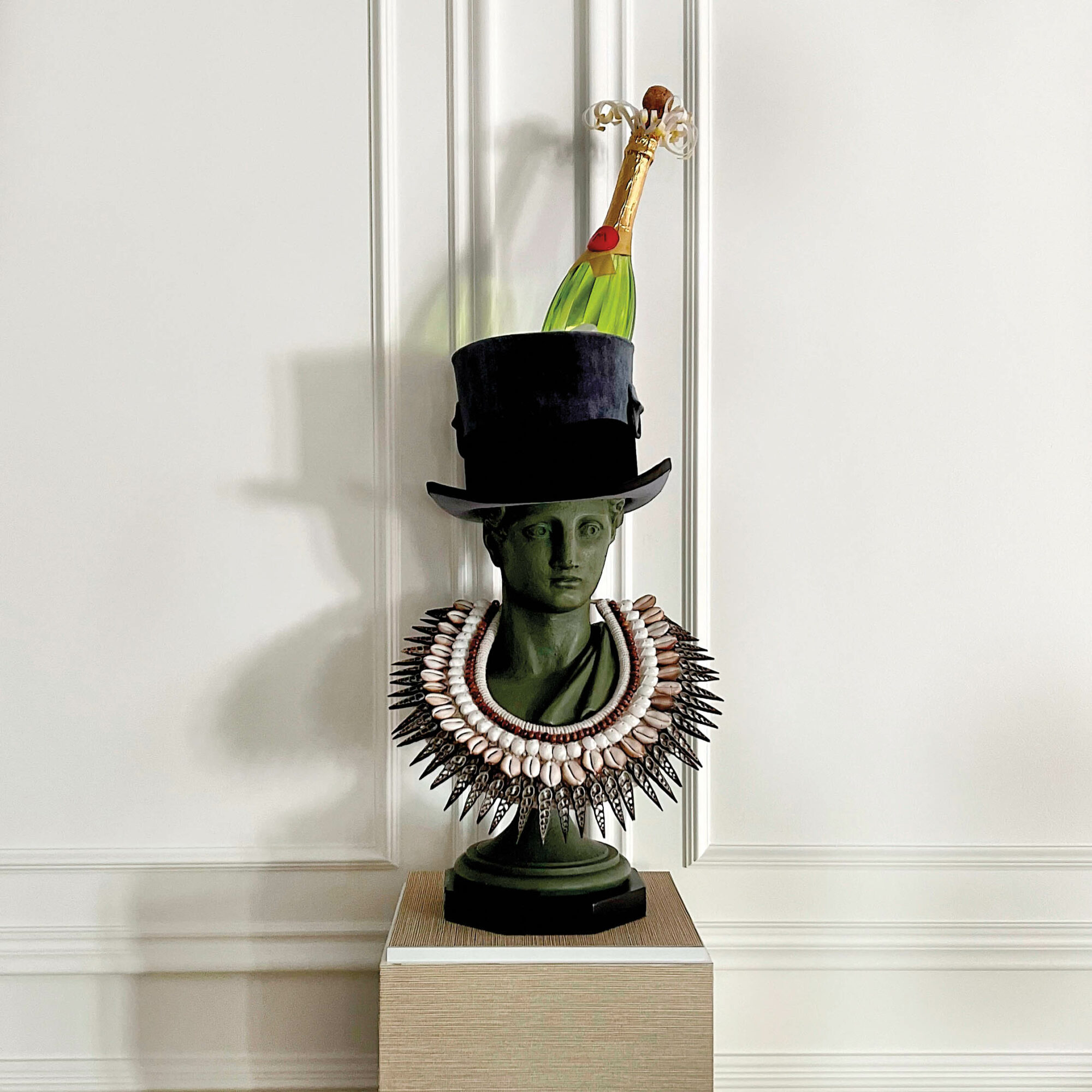 Dark green bust on a pedestal wearing a shell necklace and top hat with a champagne bottle sticking out