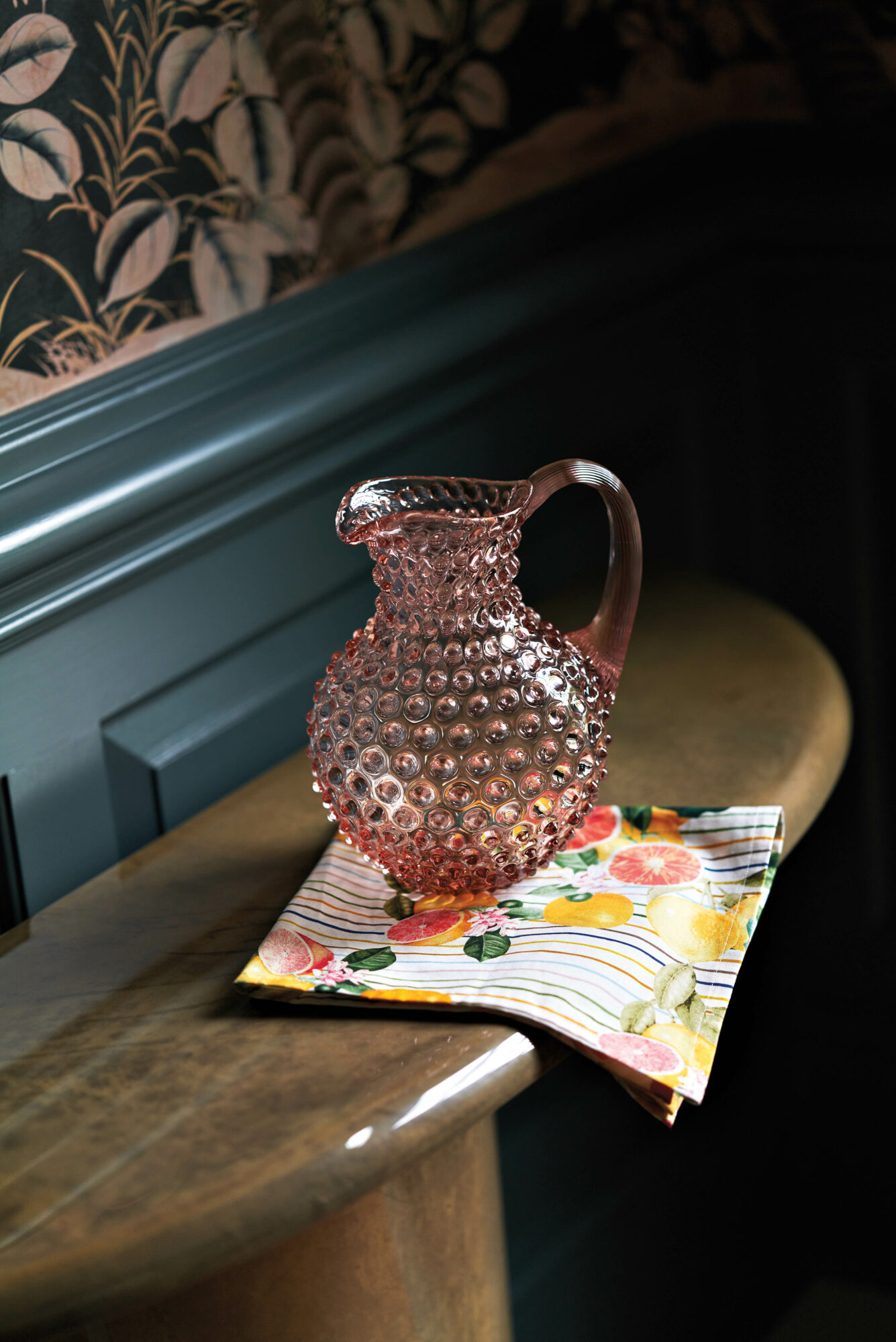 A pink-glass pitcher sits on a citrus-print napkin, by Charlotte Lucas, atop a wooden console
