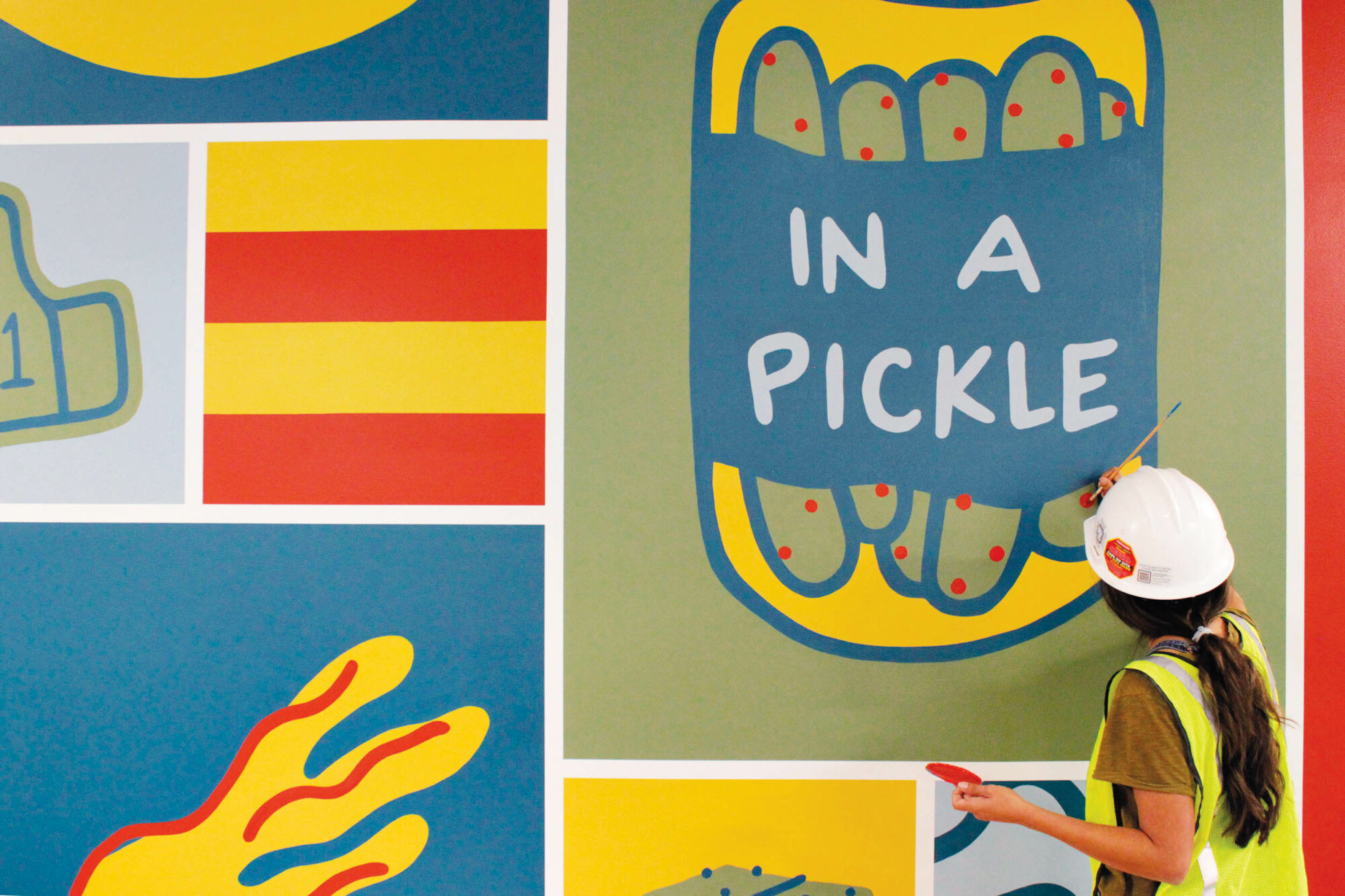 Artist Nicole Poppell painting a pickle jar as part of a larger food-themed mural