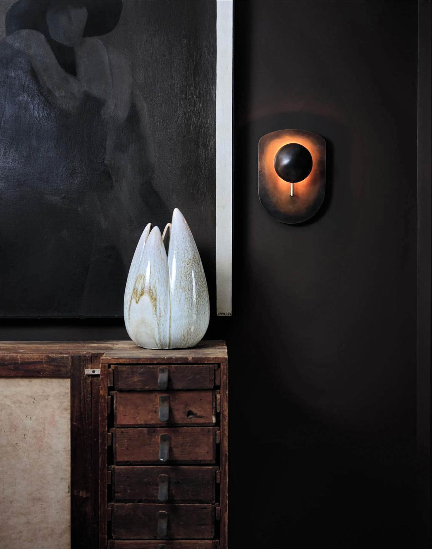 Rustic console table topped with a sculptural white vase against a black wall with a sconce by Gil Melott