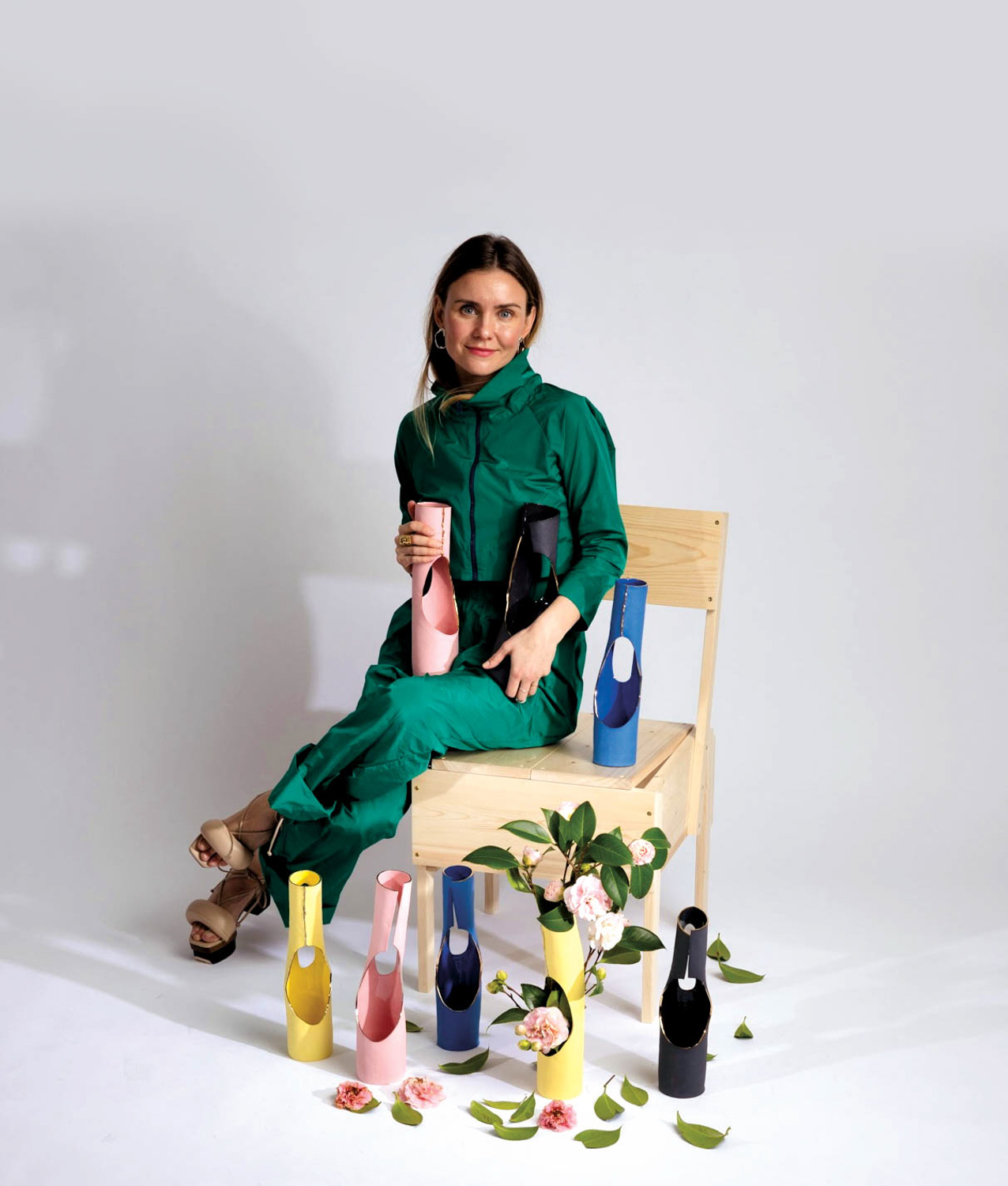 Ceramicist Evgeniya Plotnikova sitting on a wood chair with her pink, yellow, black and blue vases