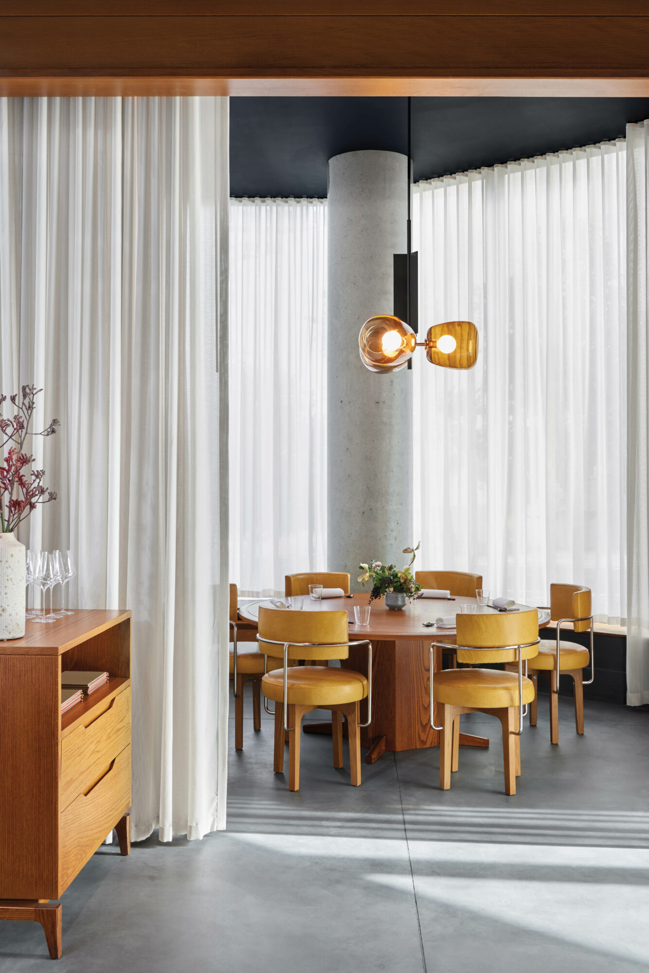 Round wooden table surrounded by wood chairs with yellow leather upholstery and sheer white curtains at Akikos