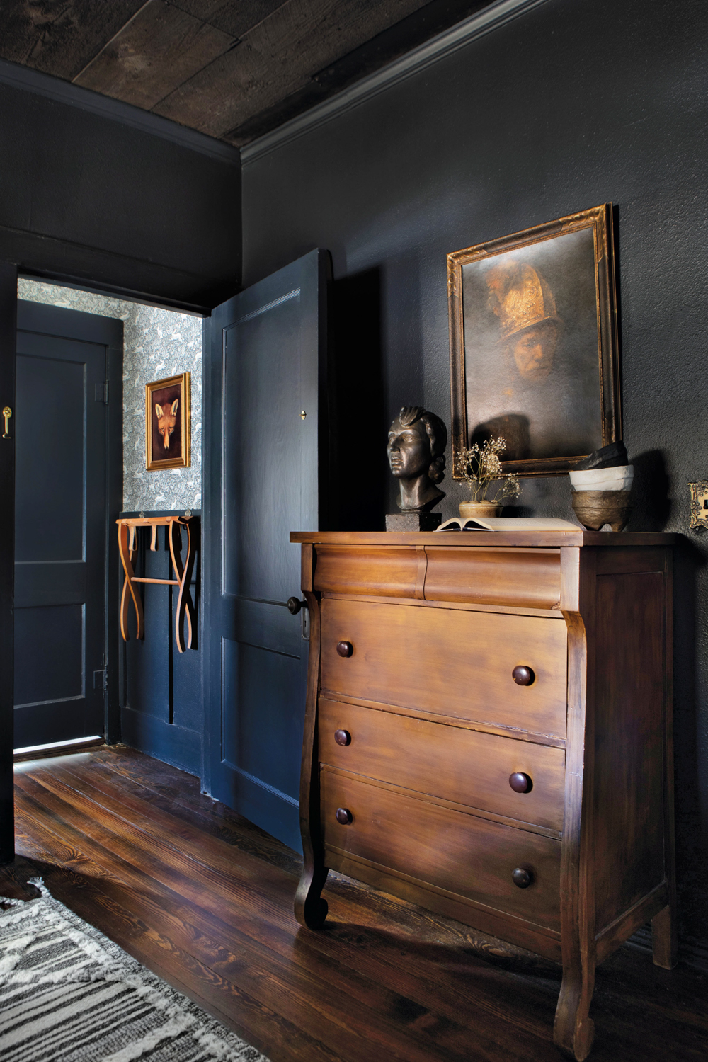 Room with painting, and a 3-drawer wooden dresser standing against midnight-blue walls and an open door