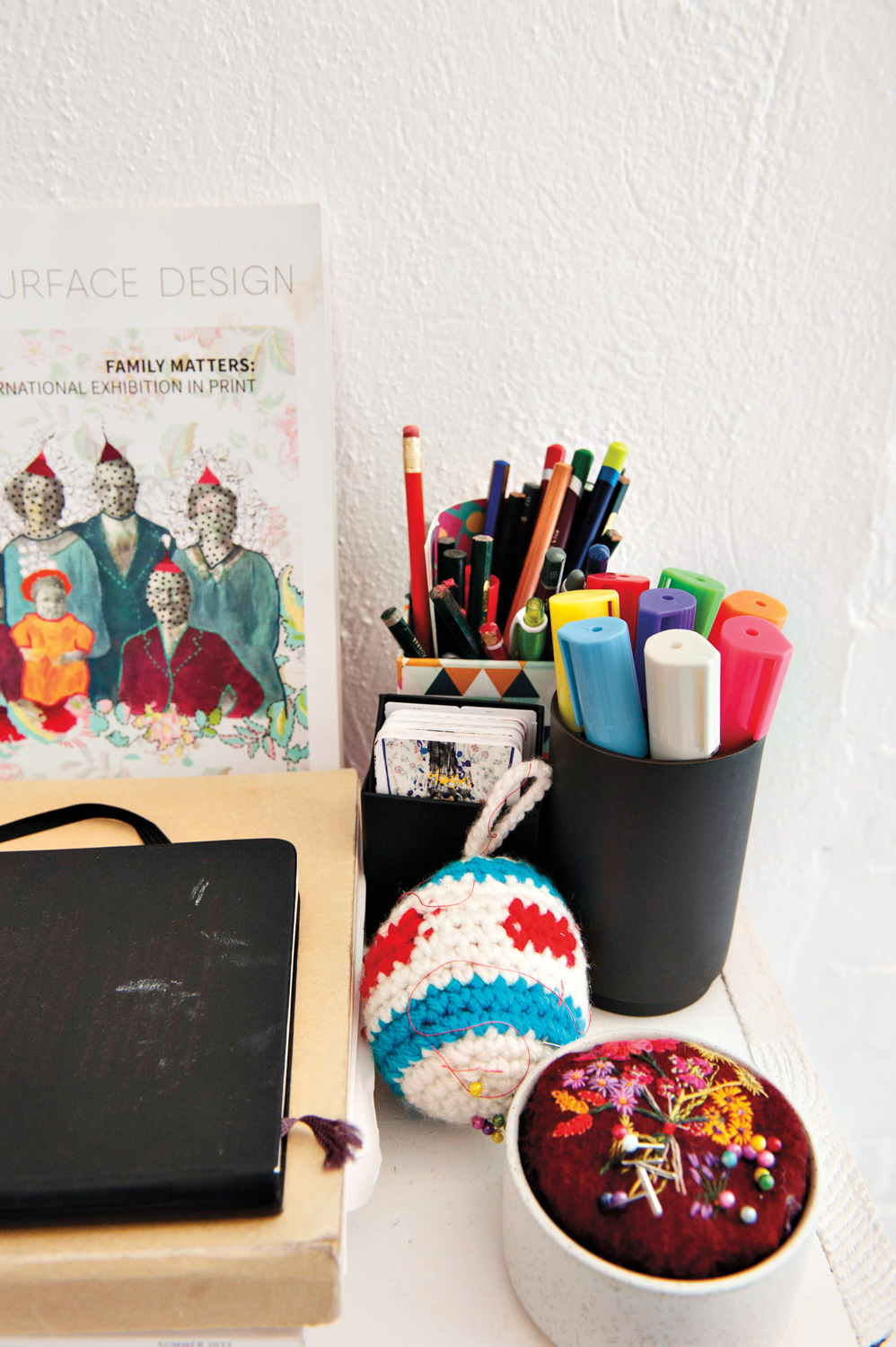 a desk with cups of markers and pencils, a pin cushion, a notebook and an exhibition catalog
