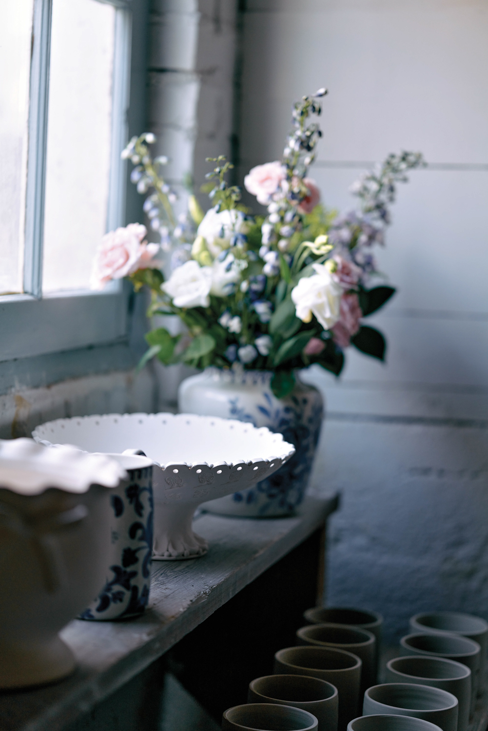 Shelf of ceramics and flowers next to a window providing dim, cool light