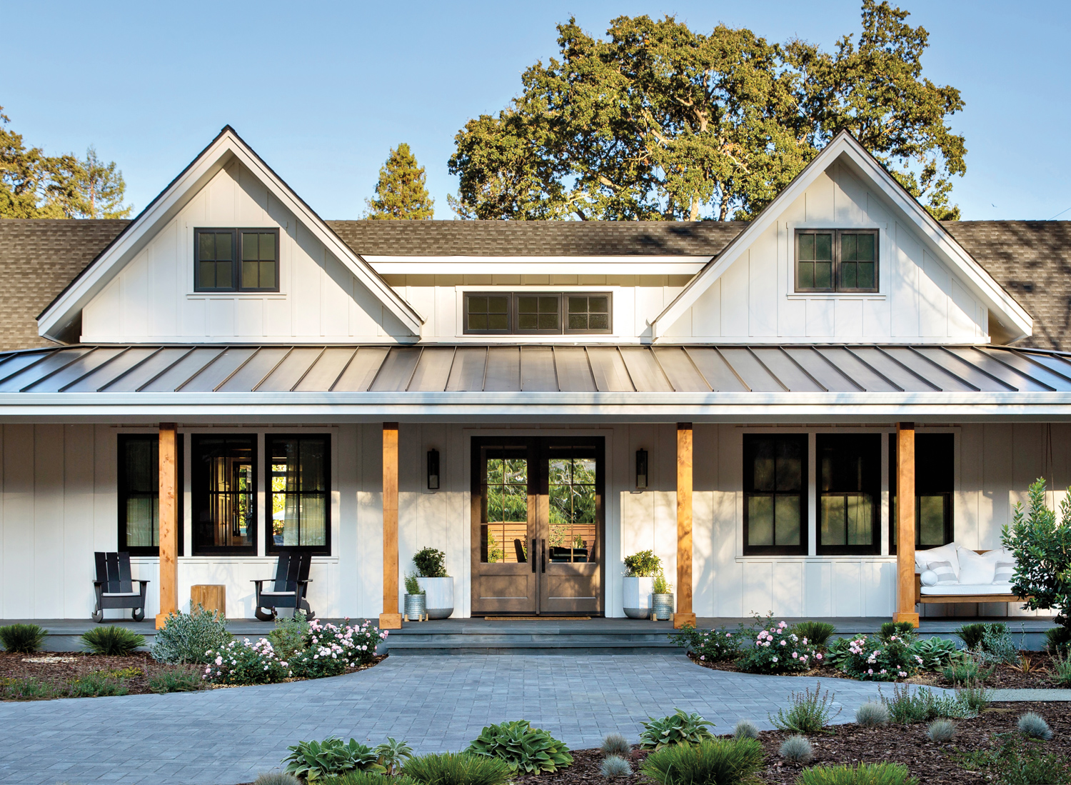 white modern farmhouse exterior with wood beams, small potted gardens and black chairs on porch
