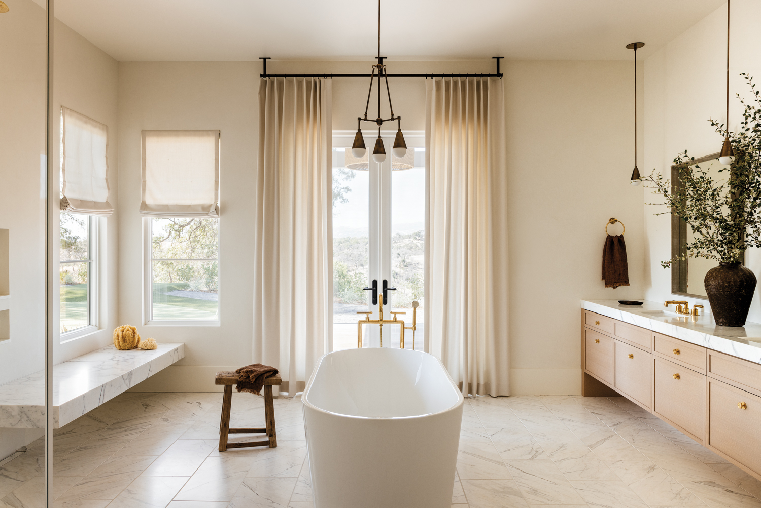bathroom tub overlooking outside space with oak cabinetry, neutral tile and brass light fixture