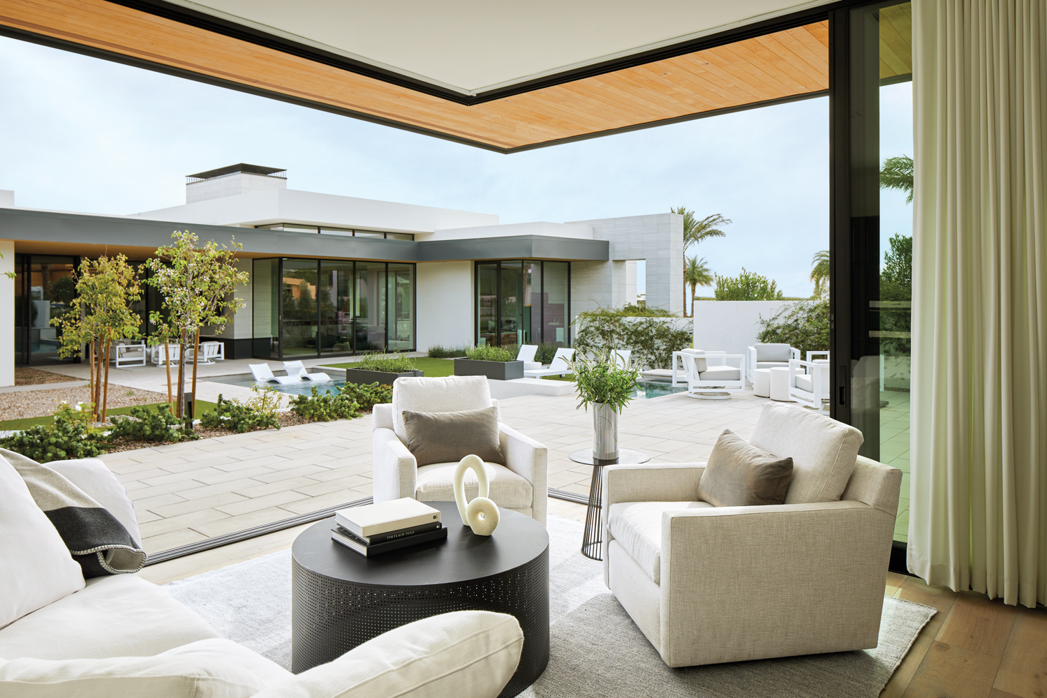 Living room with white chairs, a black coffee table and walls that open to an outdoor patio