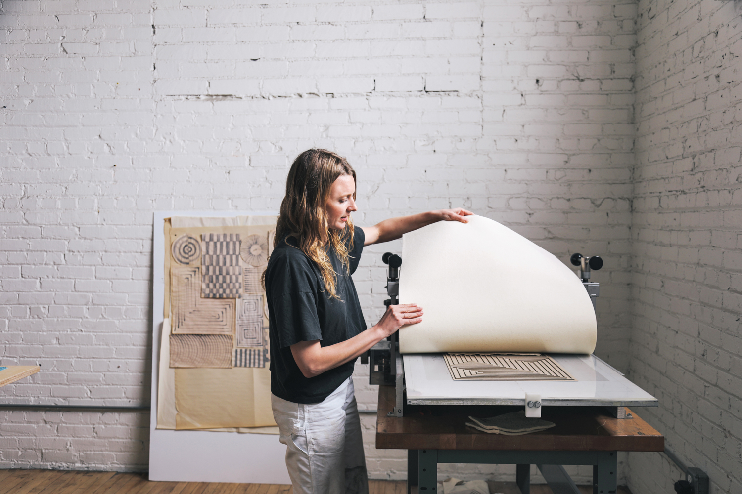artist Cheryl Humphreys stands beside a paper press in front of sketches and white brick wall