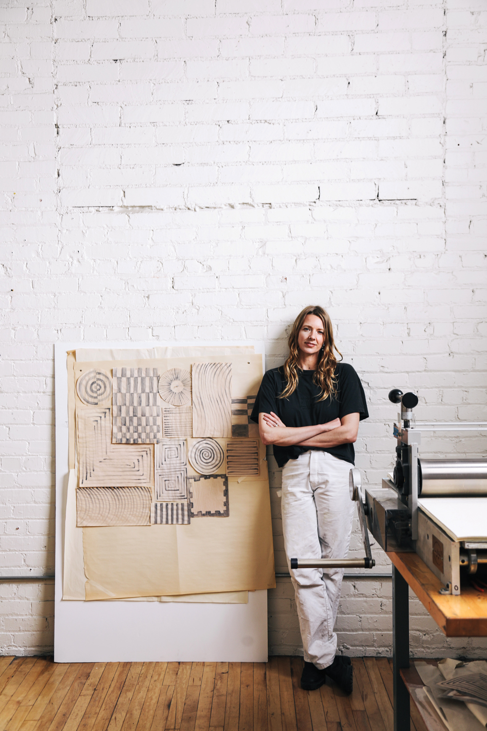 Cheryl Humphreys stands in front of a paper press and hand drawn sample works against a white brick wall