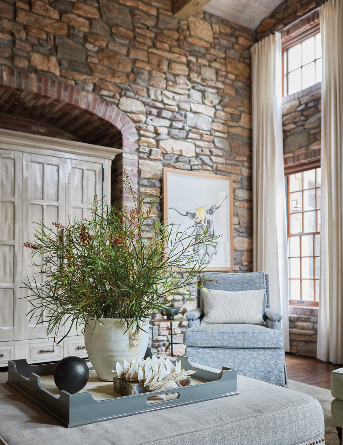 A blue armchair, gray uplhostered coffee table with a plant atop, and a tall gray wood dresser in a room with natural-stone walls.