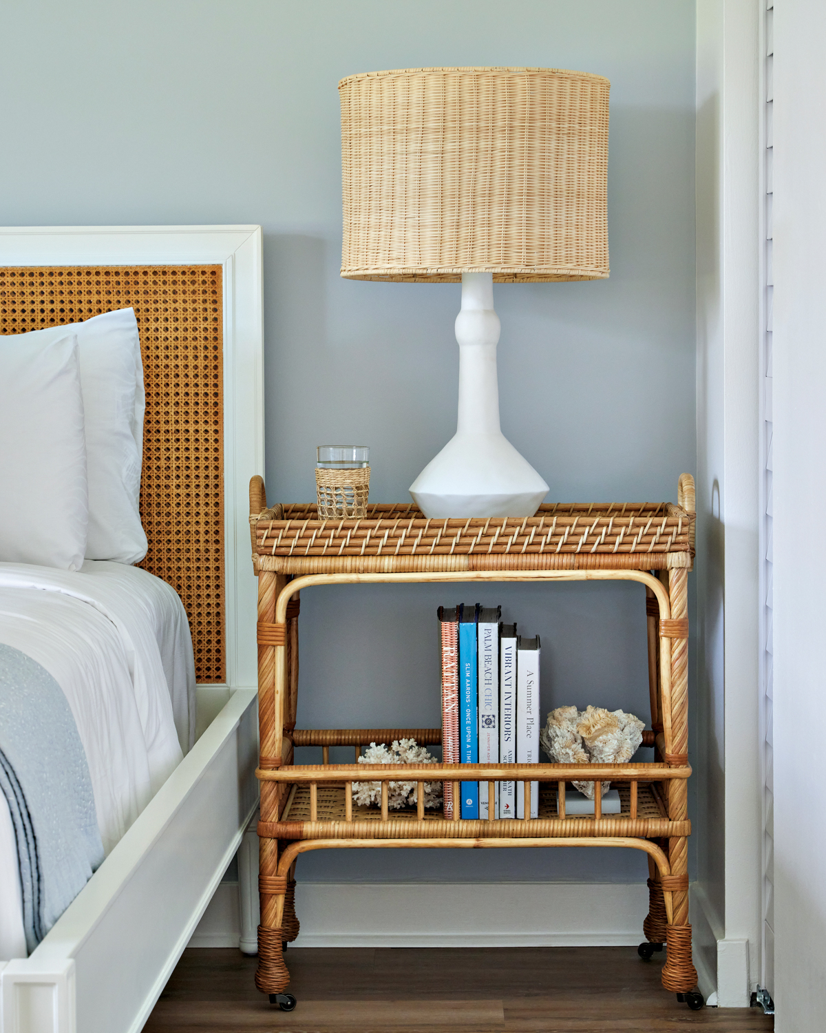Table lamp with a wicker shade on a wicker side cart next to a white cane pattern bed by Serena & Lily