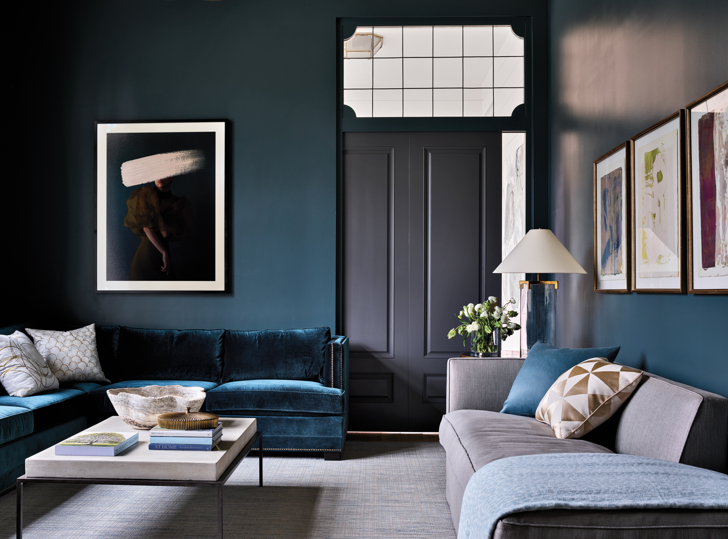 Family room with abstract artwork, velvet blue sofa, gray chaise, white table lamp and slate pocket door