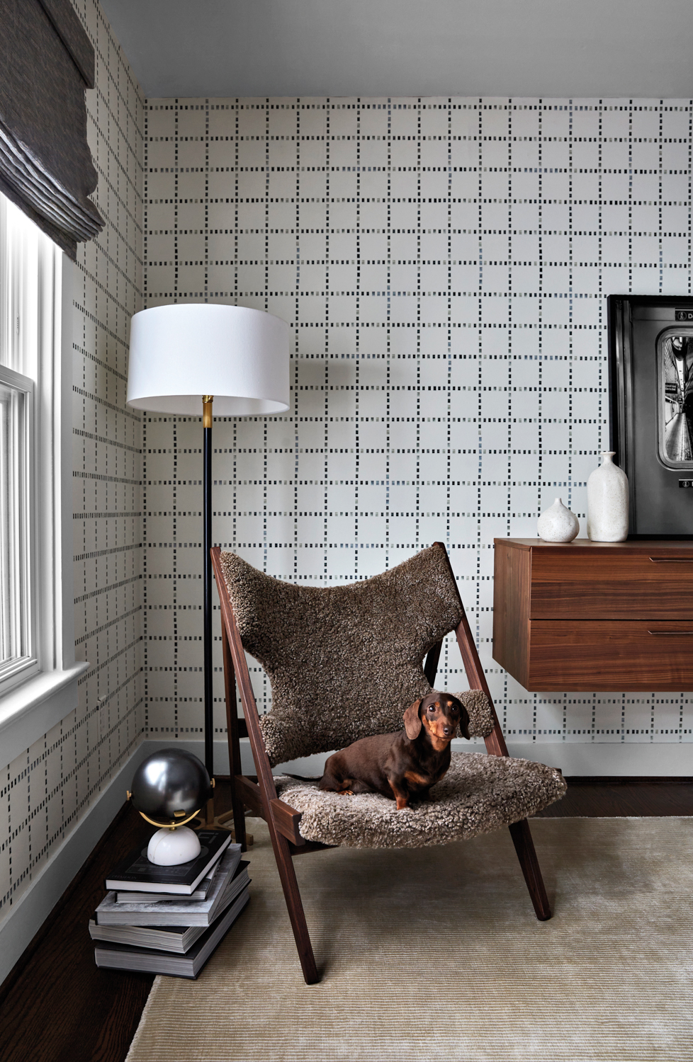 A dog sits on a chair perched in the corner in front of a floor lamp and window-pane plaid wallpapered walls