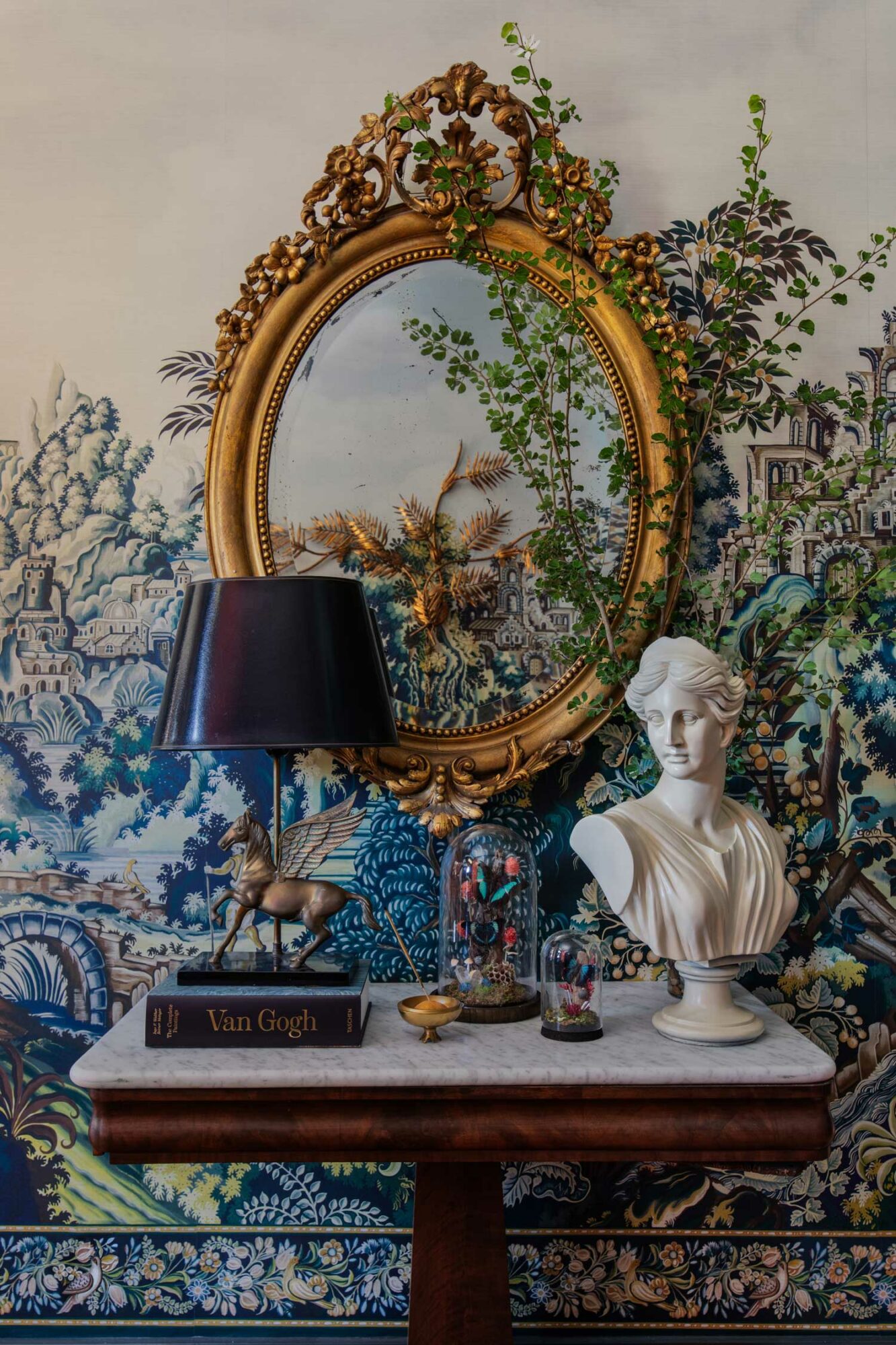 room with table, circular mirror, statue bust, and one of the 2023 wallpaper trends: verdure tapestry