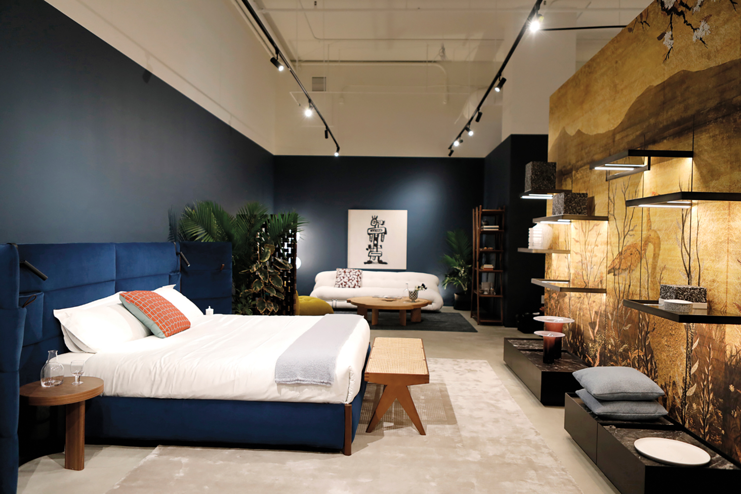 Showroom with blue upholstered bed, wood bench and side table, and white sofa beneath and black-and-white artwork