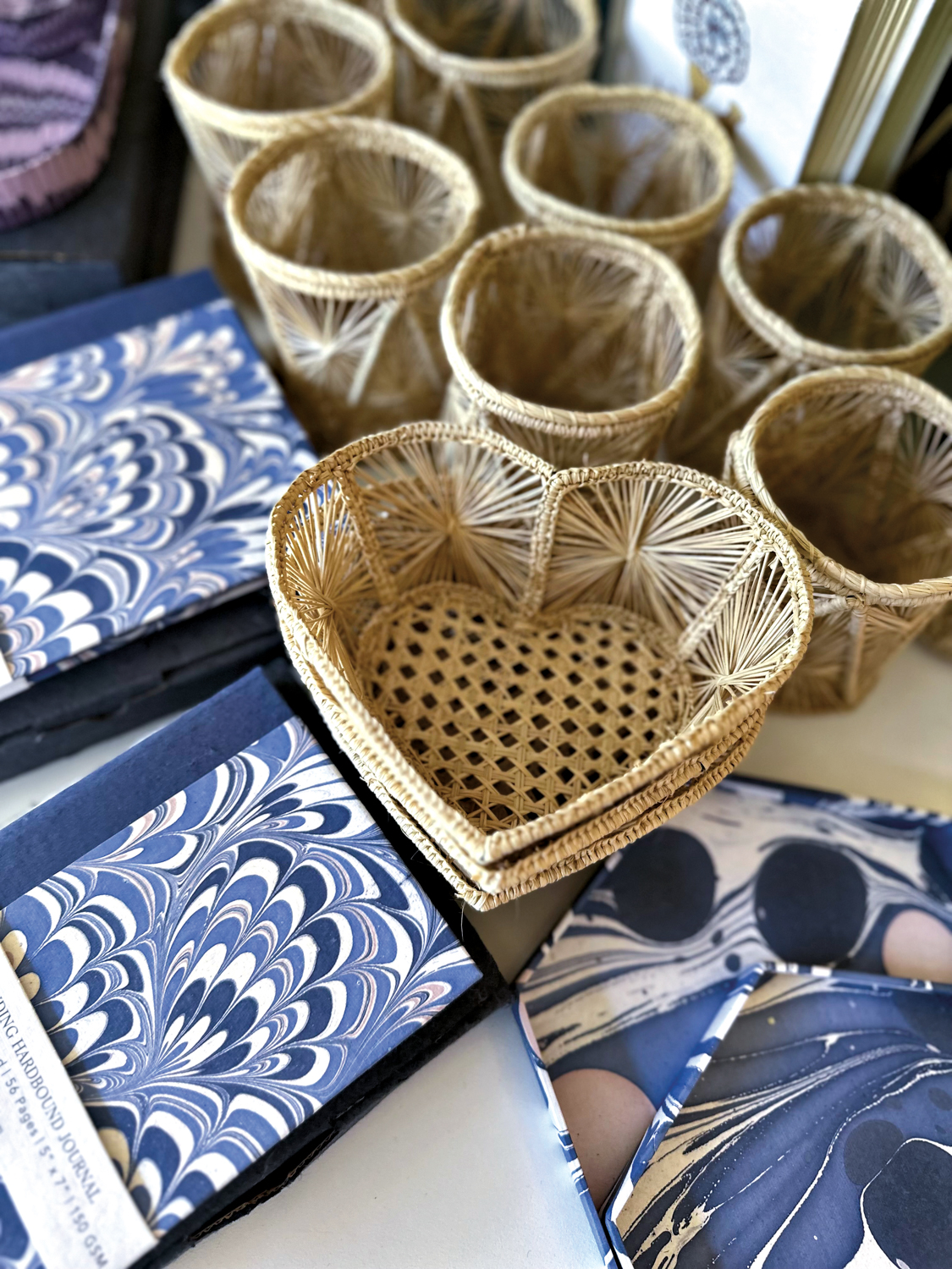 Small, woven heart-shaped basket and blue notebooks in shop by Table topped with greenery, notebooks and colorful ceramics in shop by Josephine Fisher Freckmann