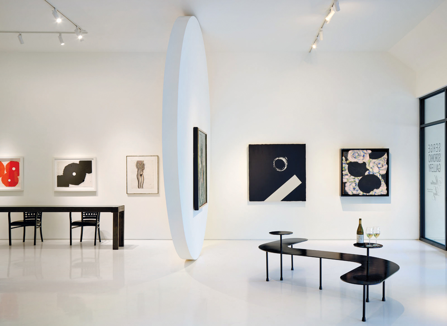 White-walled gallery with black, white and red artwork on the walls and curved black metal benches.