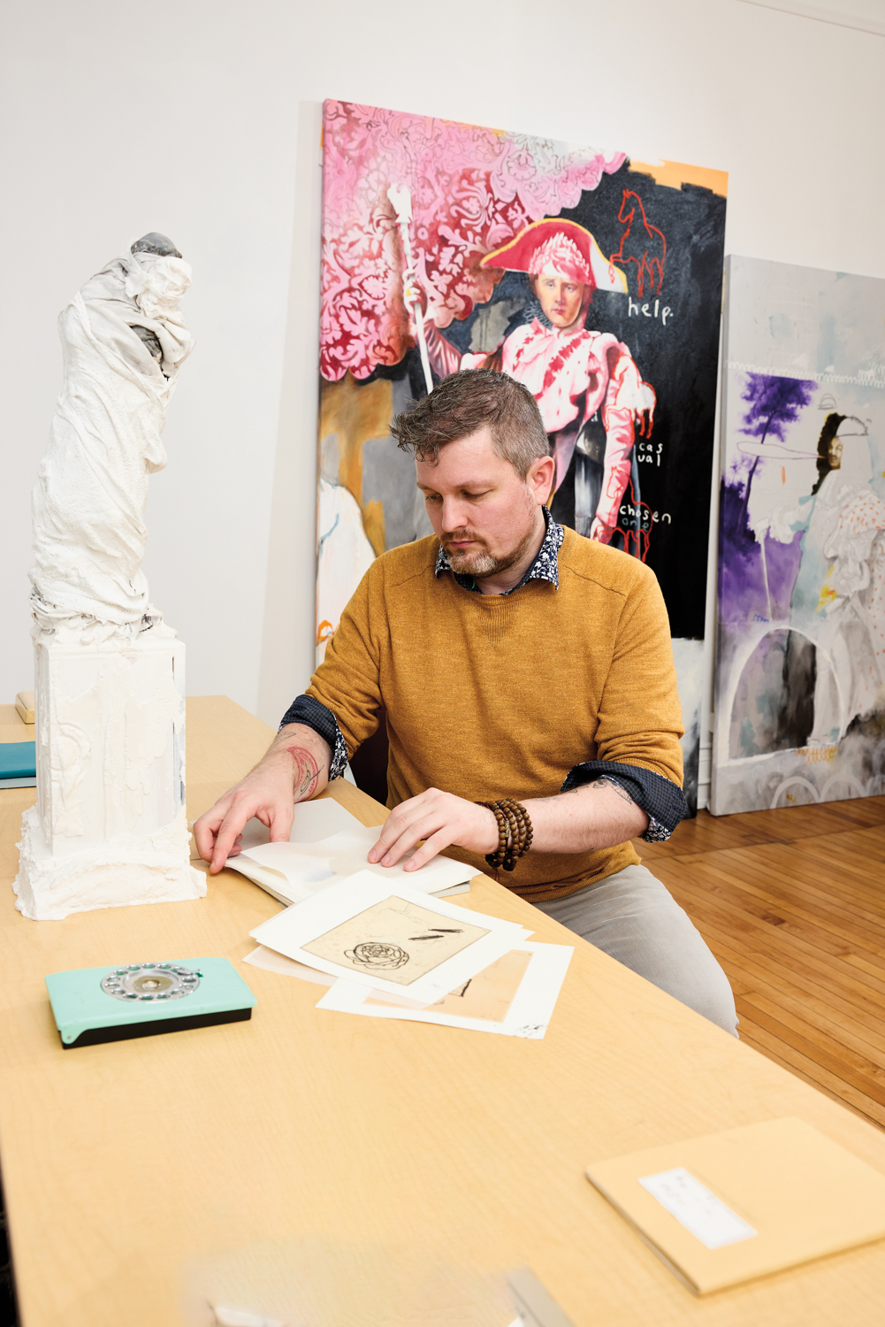 Artist Matt Bodett flipping through papers with two historically inspired paintings behind him