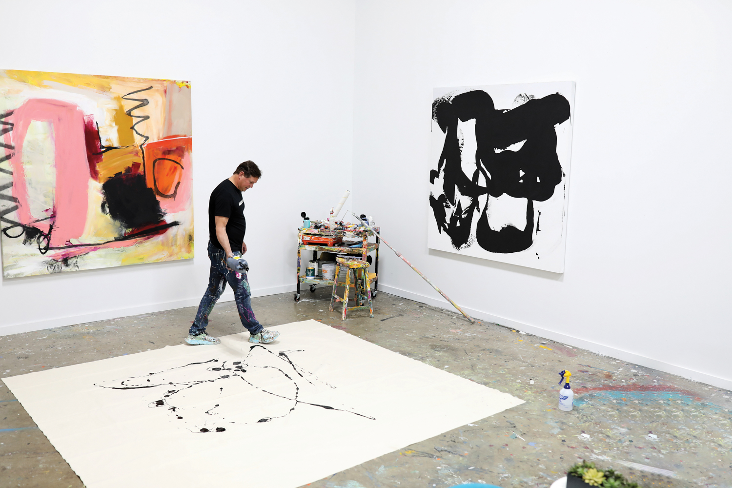 Will Day walking around a canvas spread out on the floor, dropping black paint on it