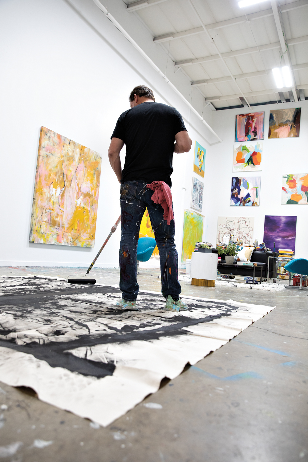 artist standing and using a broom to move paint around on a canvas lying on the floor