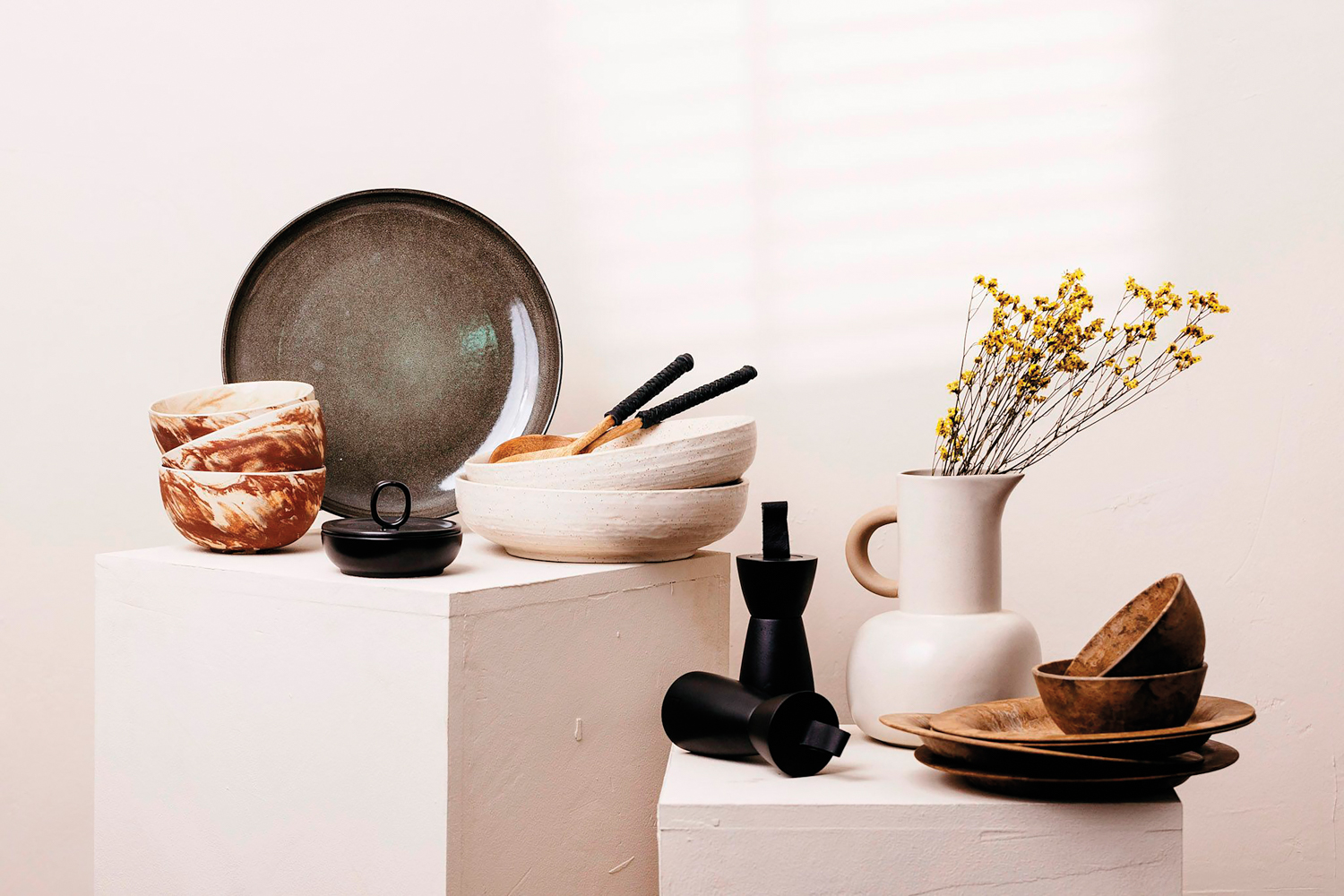 Black and cream-colored ceramic dishware and wood bowls on white plinths by Kameran Schaffner
