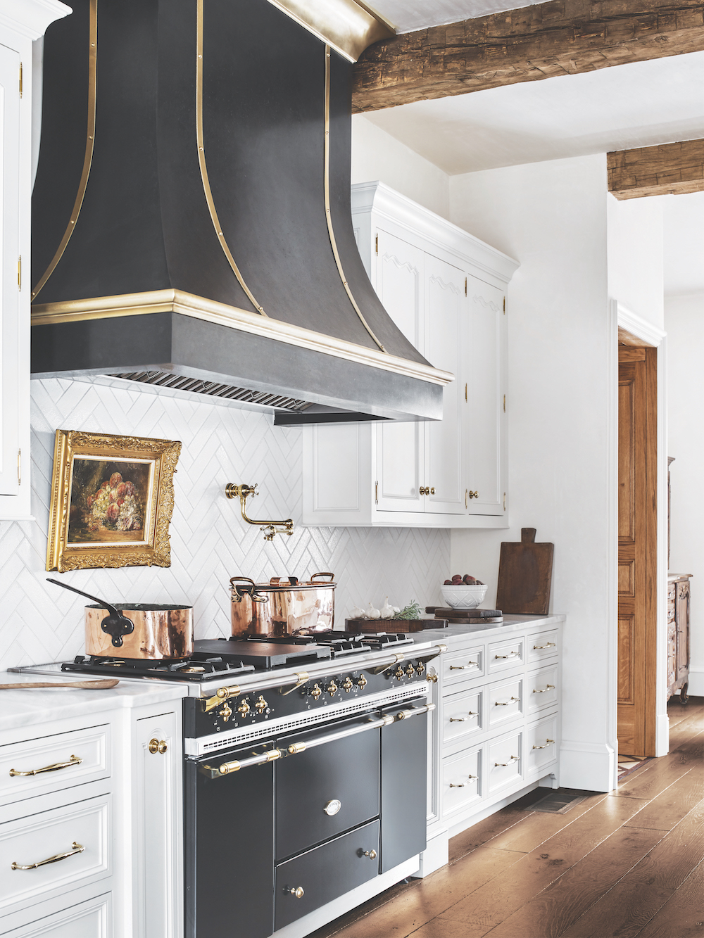 The Aurore Range Hood by Francois & Co in open kitchen with white kitchen cabinets and wood beams