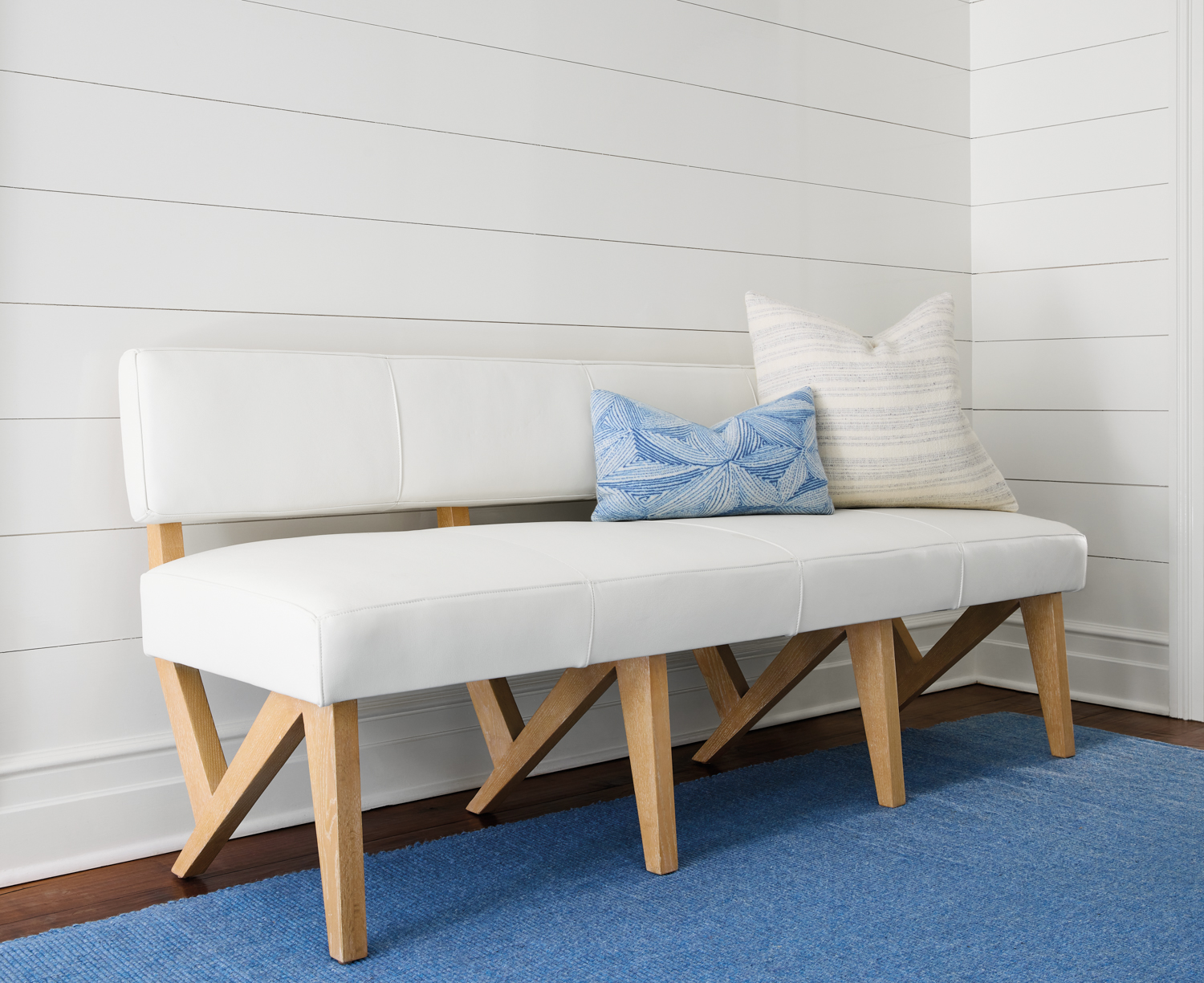 Bench with white upholstery and light wood frame topped and with blue and white pillows on a blue rug.