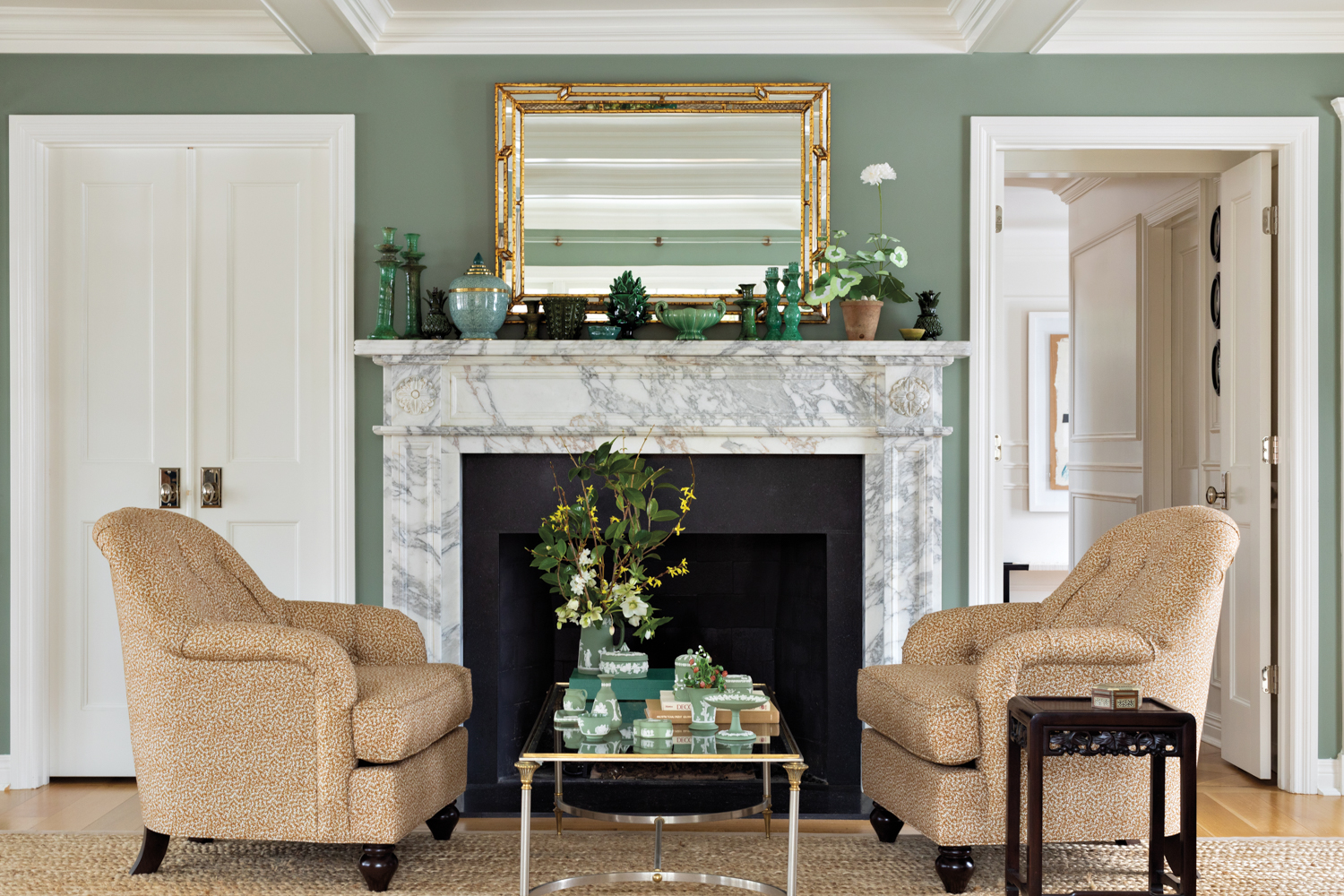 Two armchairs facing each other with a glass-and-metal coffee table between, in front of a marble fireplace and green wall.