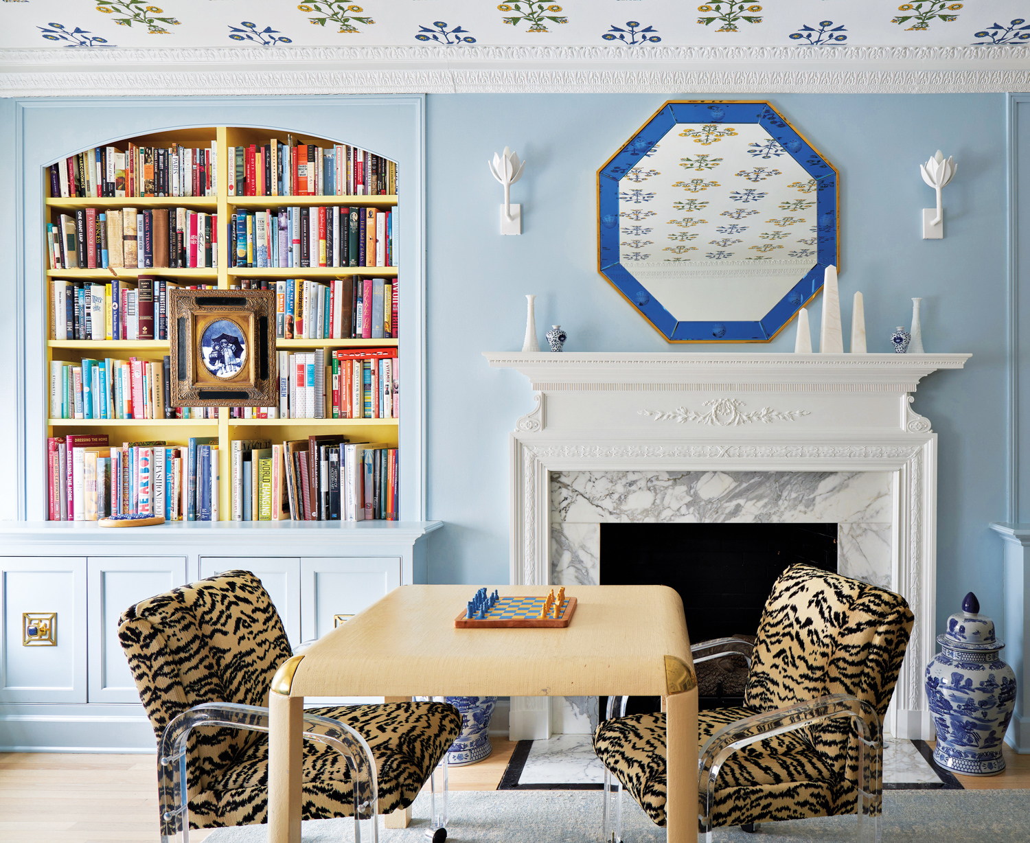 Leopard-print armchairs and a wood table in front of a marble fireplace and bookshelves by Jasmin Reese