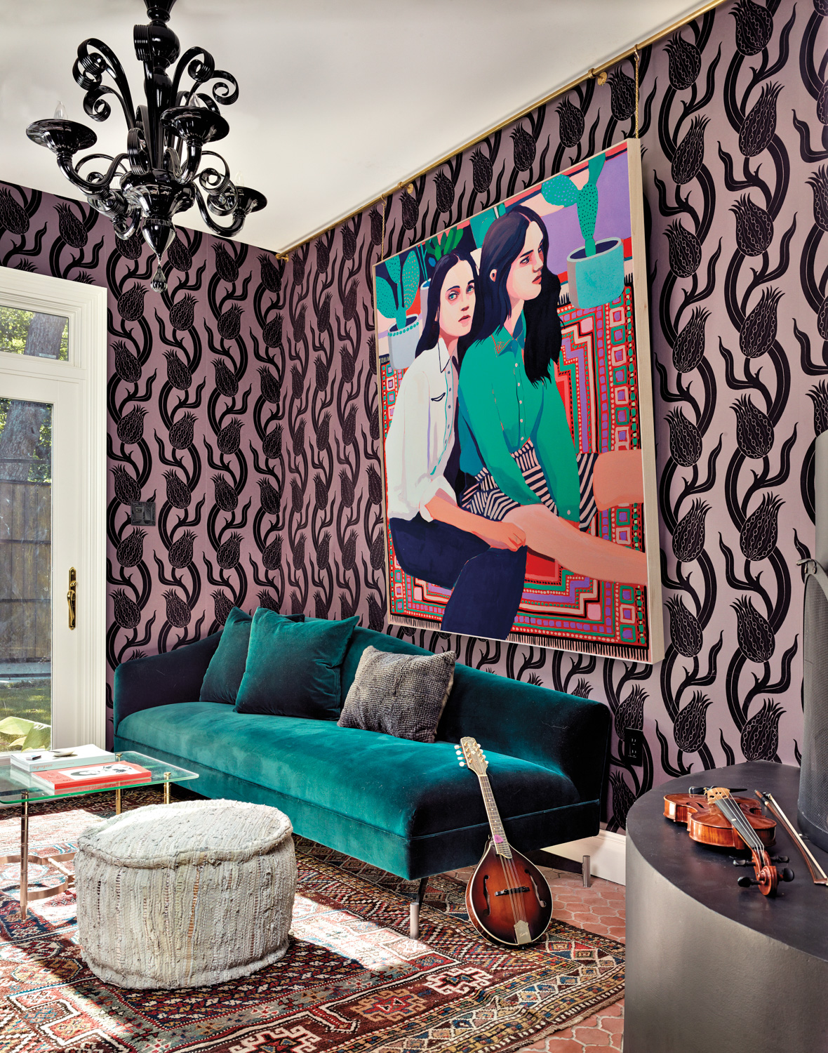 Living room with a large-scale painting of 2 women against boldly patterned wallpaper above a teal-velvet couch.