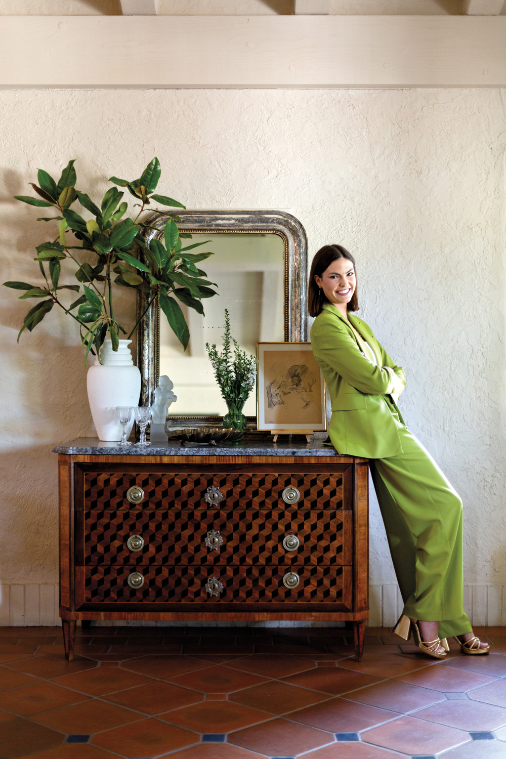 Lily Barfield in a bright green suit leaning against a dark wood sideboard.