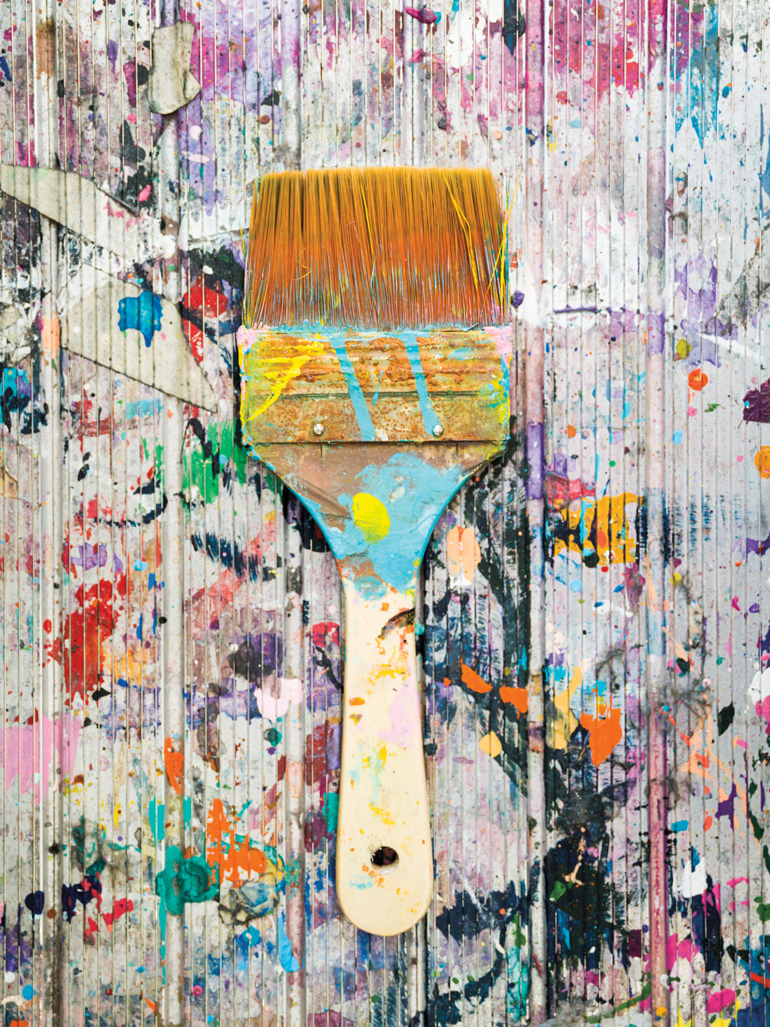 wide paintbrush covered in stripes of paint, against a paint splattered striped surface