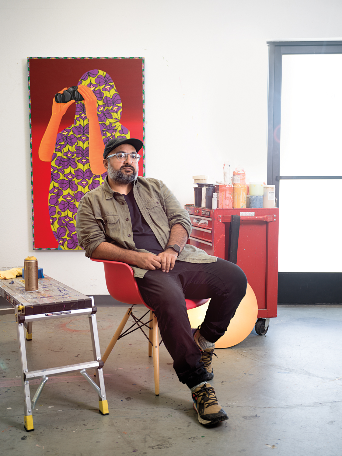 Amir H. Fallah wearing a baseball cap and glasses seated in front of painting