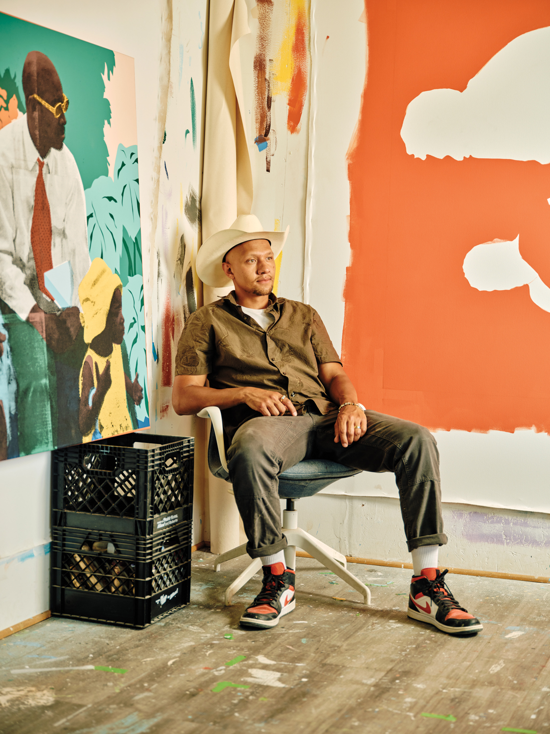 Kristopher Wright wearing cowboy hat sitting in corner between unfinished paintings on walls