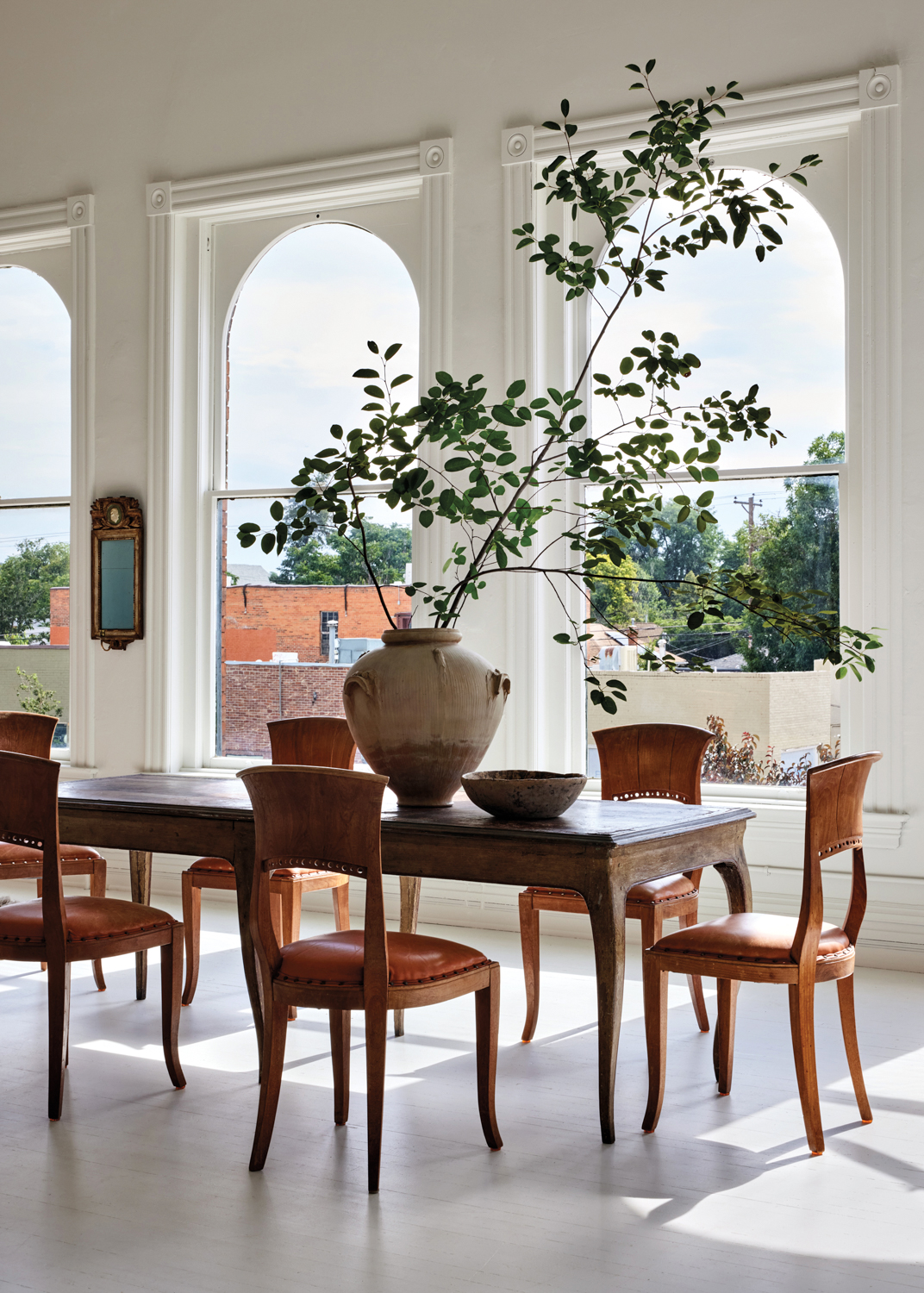 Dark-wood vintage dining table circle by wood-and-leather dining chairs, with tall branches in a vessel on top