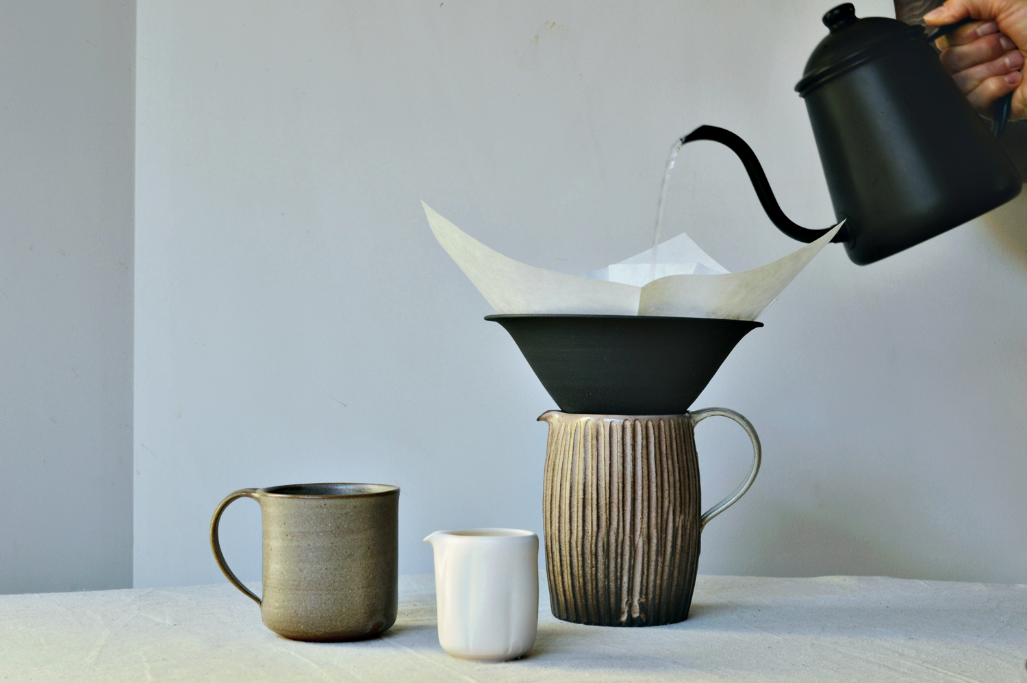 Water being poured from a black kettle into a ceramic pitcher by Kazu Oba, with two ceramic cups beside it