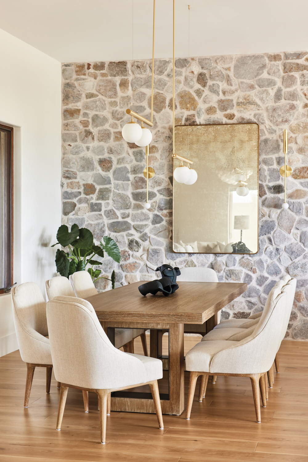 Dining room with cream-colored upholstered chairs around a wood table in front of a stone wall by Eva Higby