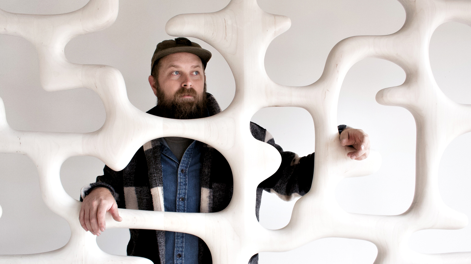 Casey Johnson stands behind a wooden sculptural wall painted white. 