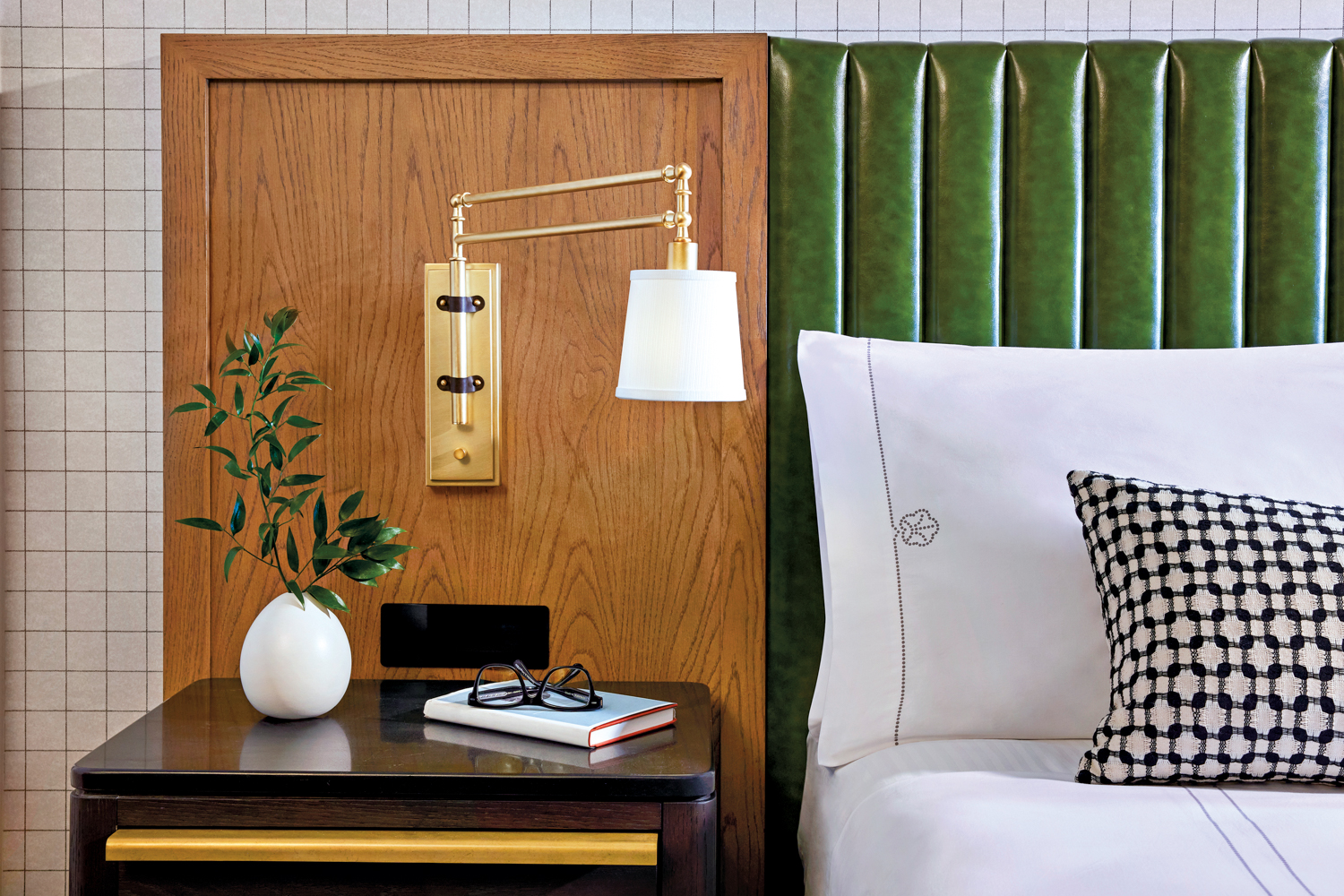 A headboard of green leather and wood backs a bed and nightstand with a sconce