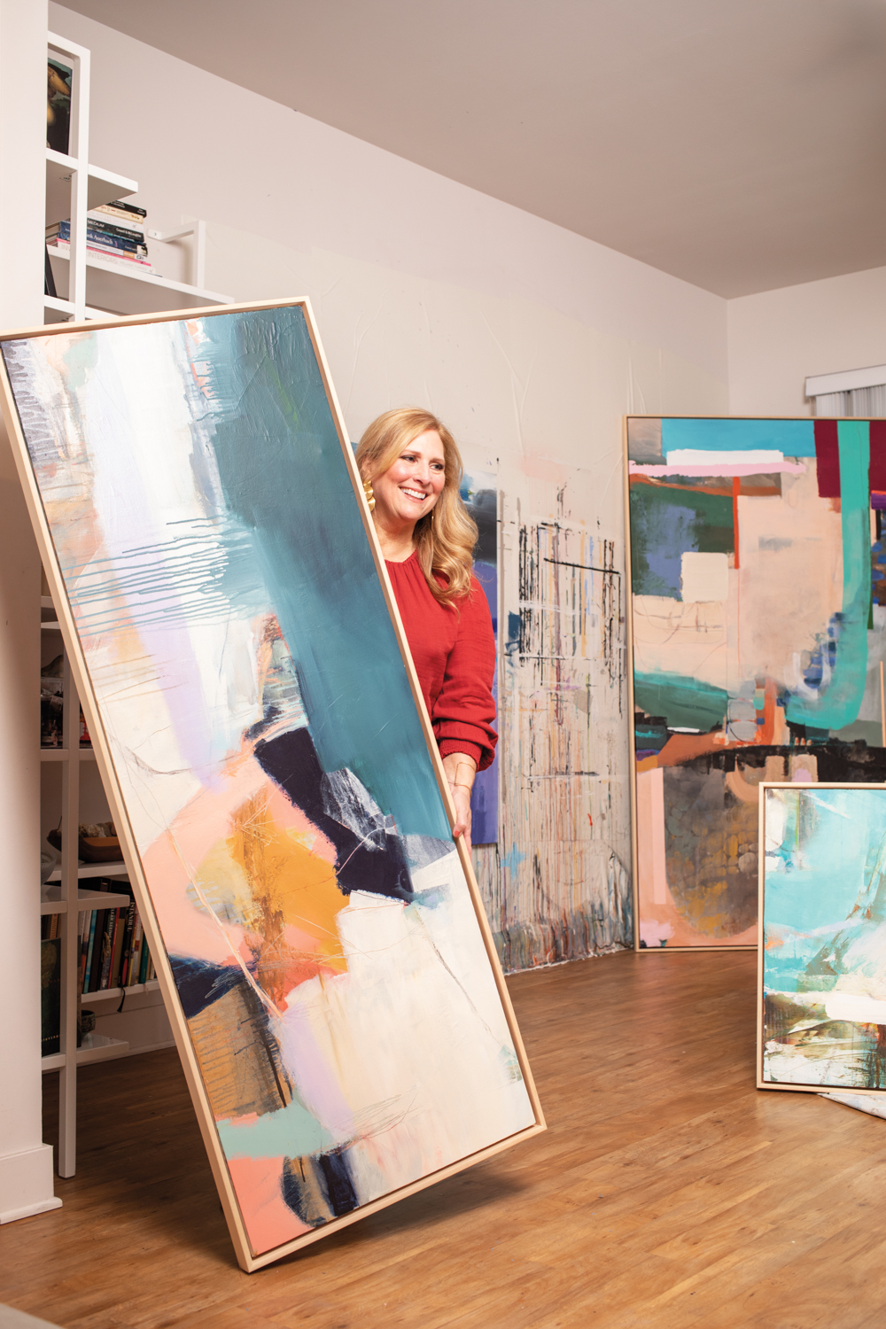 Joan Maureen Collins holding a large abstract painting with multiple paintings behind her