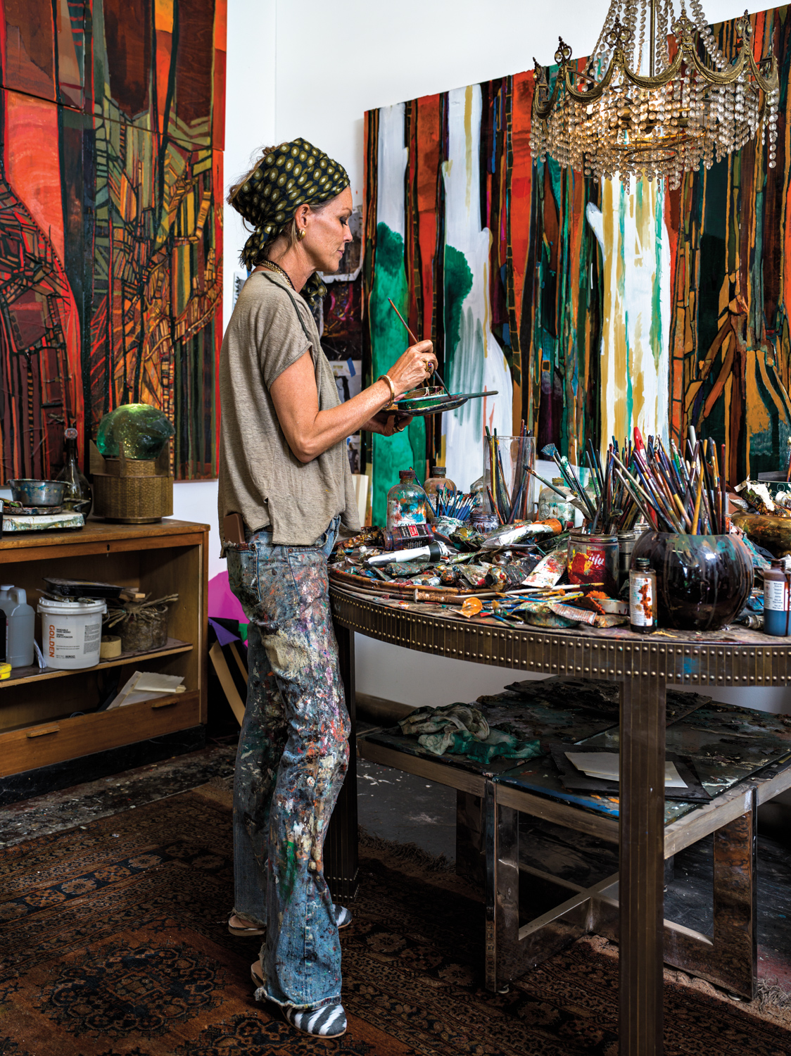 woman with headscarf standing in art studio mixing paint over a table strewn with paint brushes and paint, with canvases in the background and a glass bead chandelier hung overhead